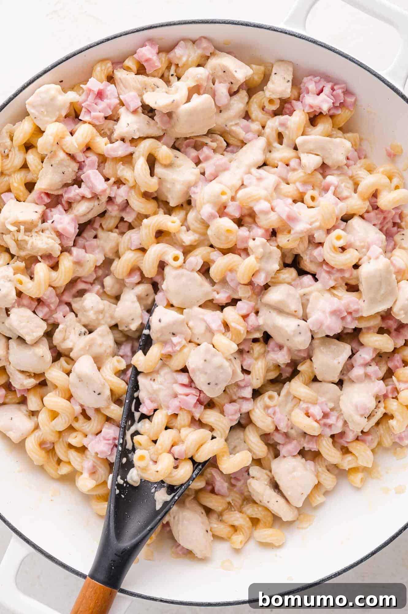 A large skillet brimming with creamy cavatappi pasta, perfectly diced chicken, and savory chopped ham, all coated in a rich white sauce, accompanied by a sleek black serving spoon.