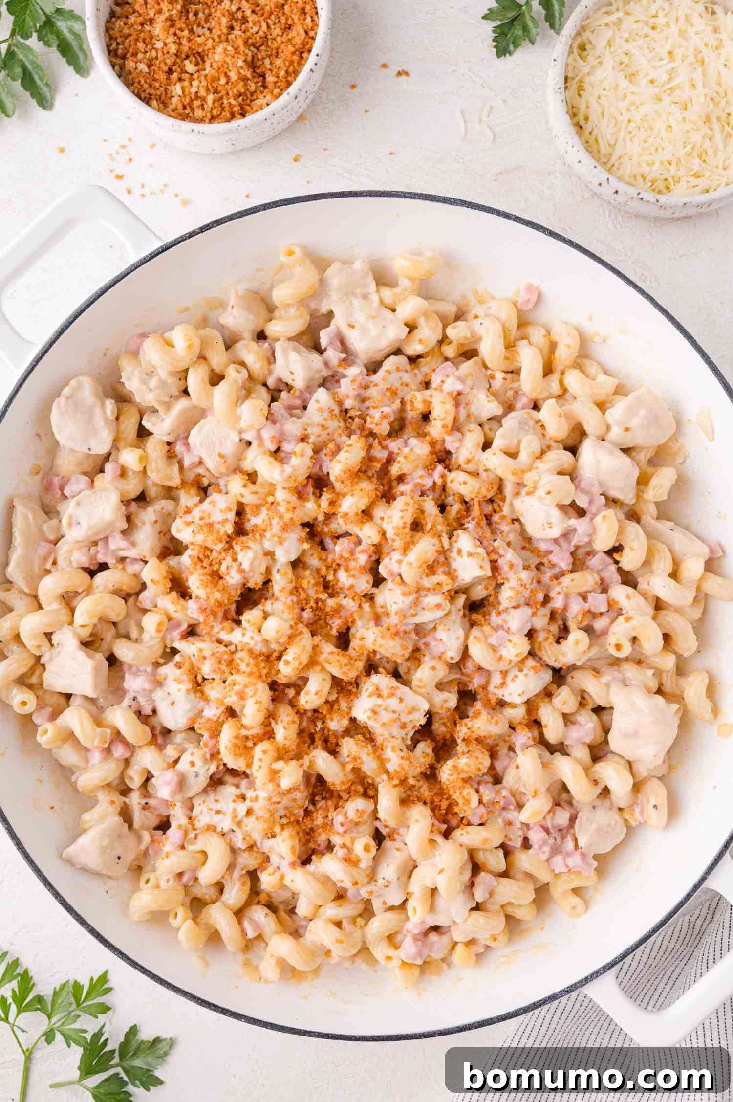 A pristine white skillet showcasing a harmonious blend of creamy pasta, tender diced chicken, and savory ham, beautifully topped with a generous sprinkle of golden toasted breadcrumbs. Nearby, small bowls hold grated cheese and additional breadcrumbs on a light surface.
