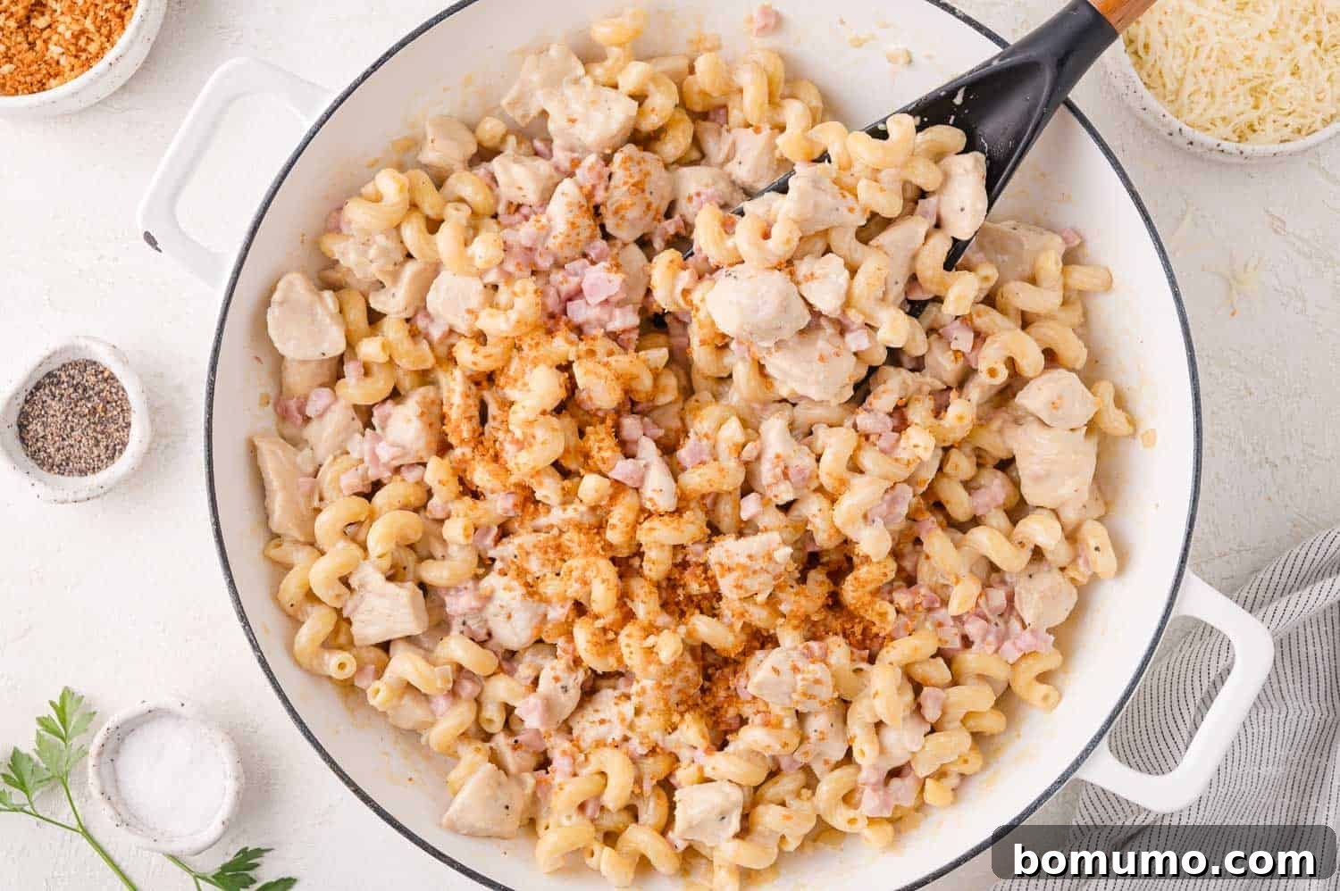 A large white skillet generously filled with creamy cavatappi pasta, succulent diced chicken, savory bacon bits, and a rich, velvety sauce, being stirred gently with a black serving spoon.
