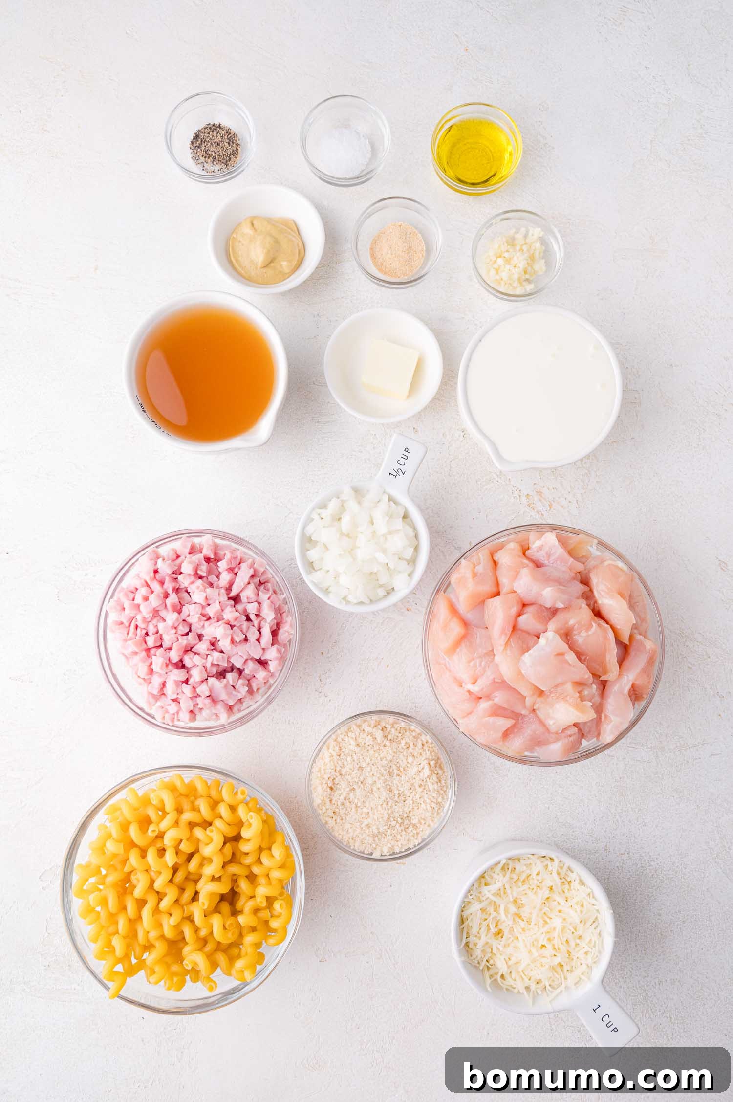 All the fresh ingredients for a delightful Chicken Cordon Bleu Pasta organized neatly on a white surface: diced chicken, spiral pasta, chopped ham, onions, broth, cream, Swiss cheese, Panko breadcrumbs, and essential seasonings.