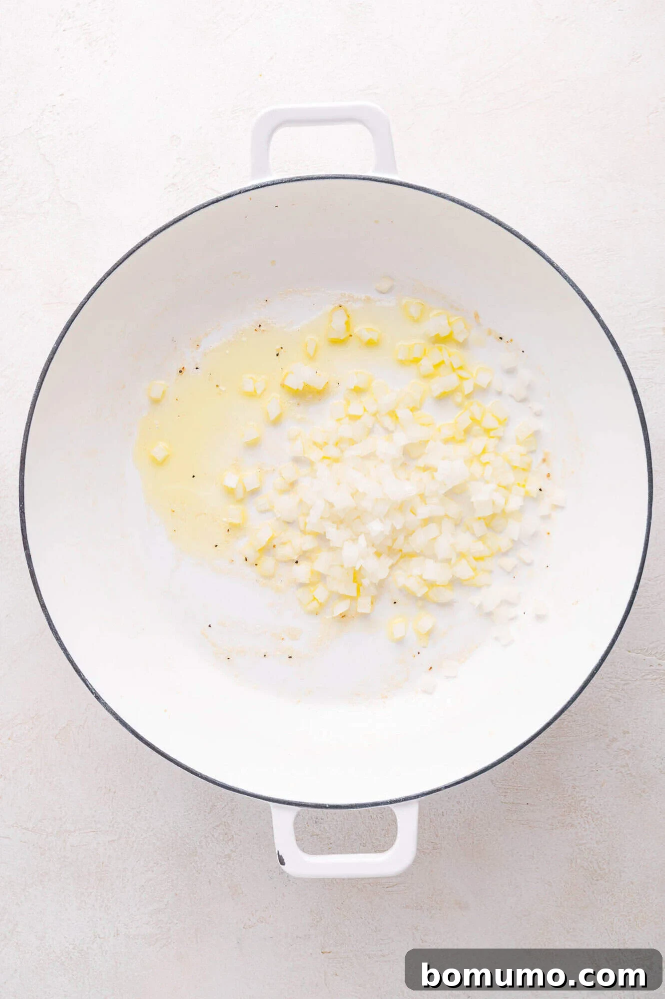 Finely diced onions gently sautéing in shimmering olive oil within a pristine white enameled pan, resting on a light-colored kitchen surface.