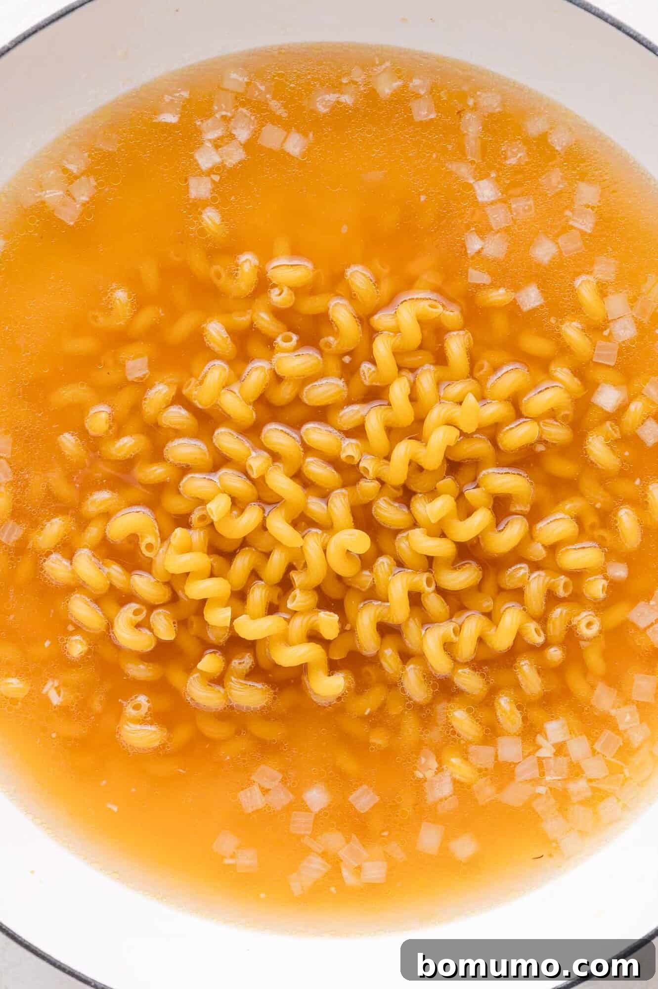A large pot filled with uncooked curly pasta, aromatic diced onions, and savory broth, ready to simmer.