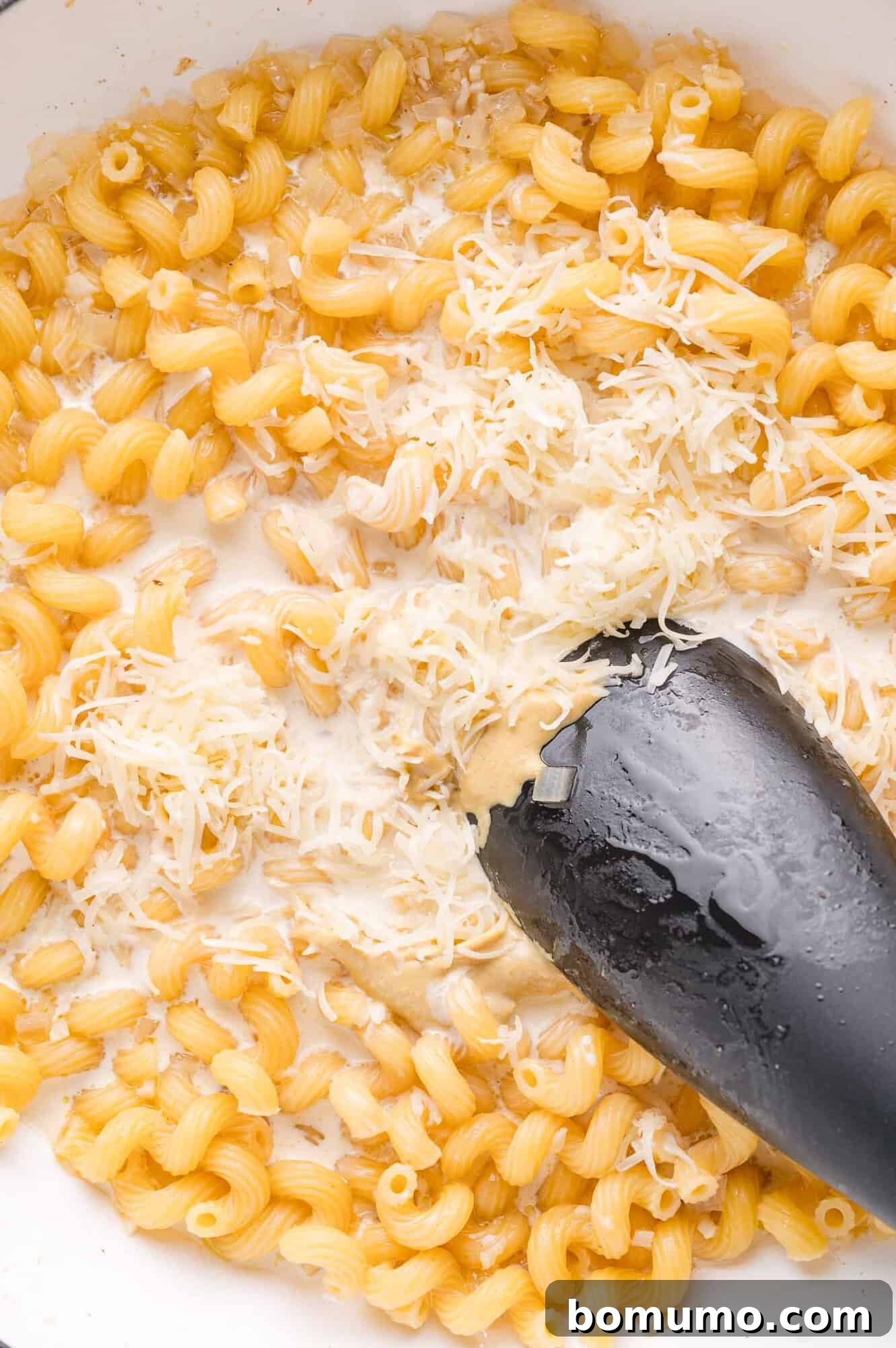 Al dente cavatappi pasta immersed in a creamy, white sauce, being stirred with a sleek black silicone spatula, with a generous sprinkle of shredded cheese melting into the mixture.