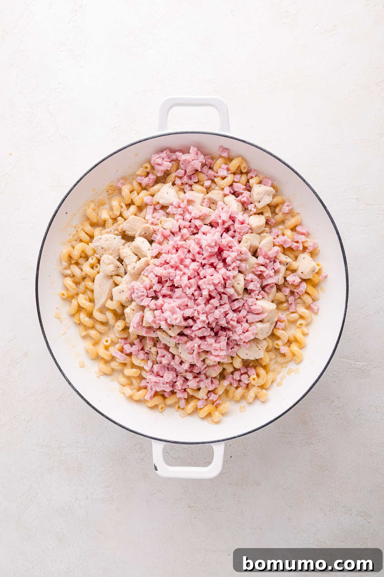 A white pot containing a harmonious blend of cooked pasta, tender diced chicken, and savory chopped ham, all awaiting the final creamy touch on a light surface.