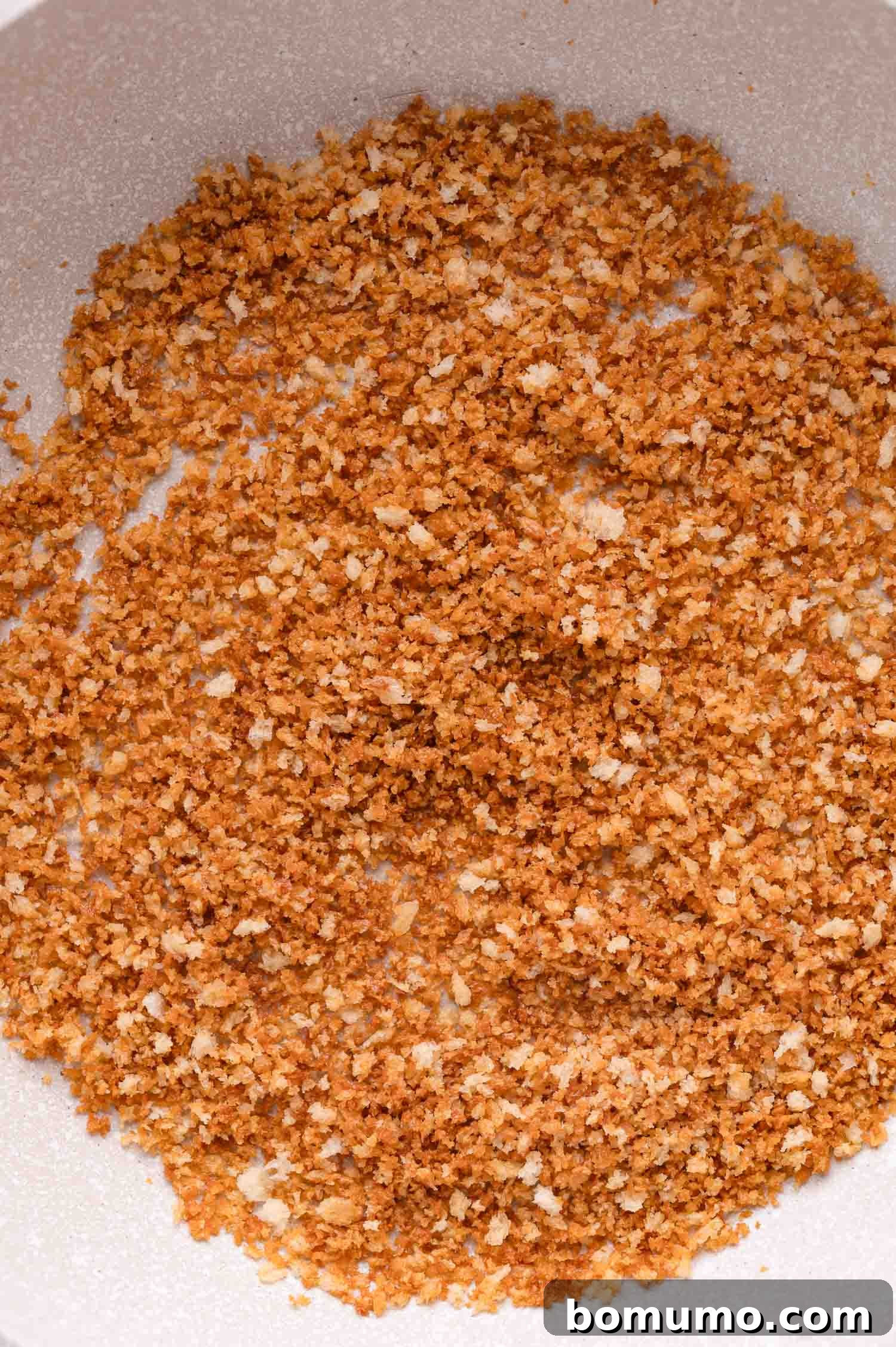 A close-up shot of perfectly golden-brown toasted breadcrumbs, evenly scattered across the bottom of a pristine white pan, ready to be used as a topping.