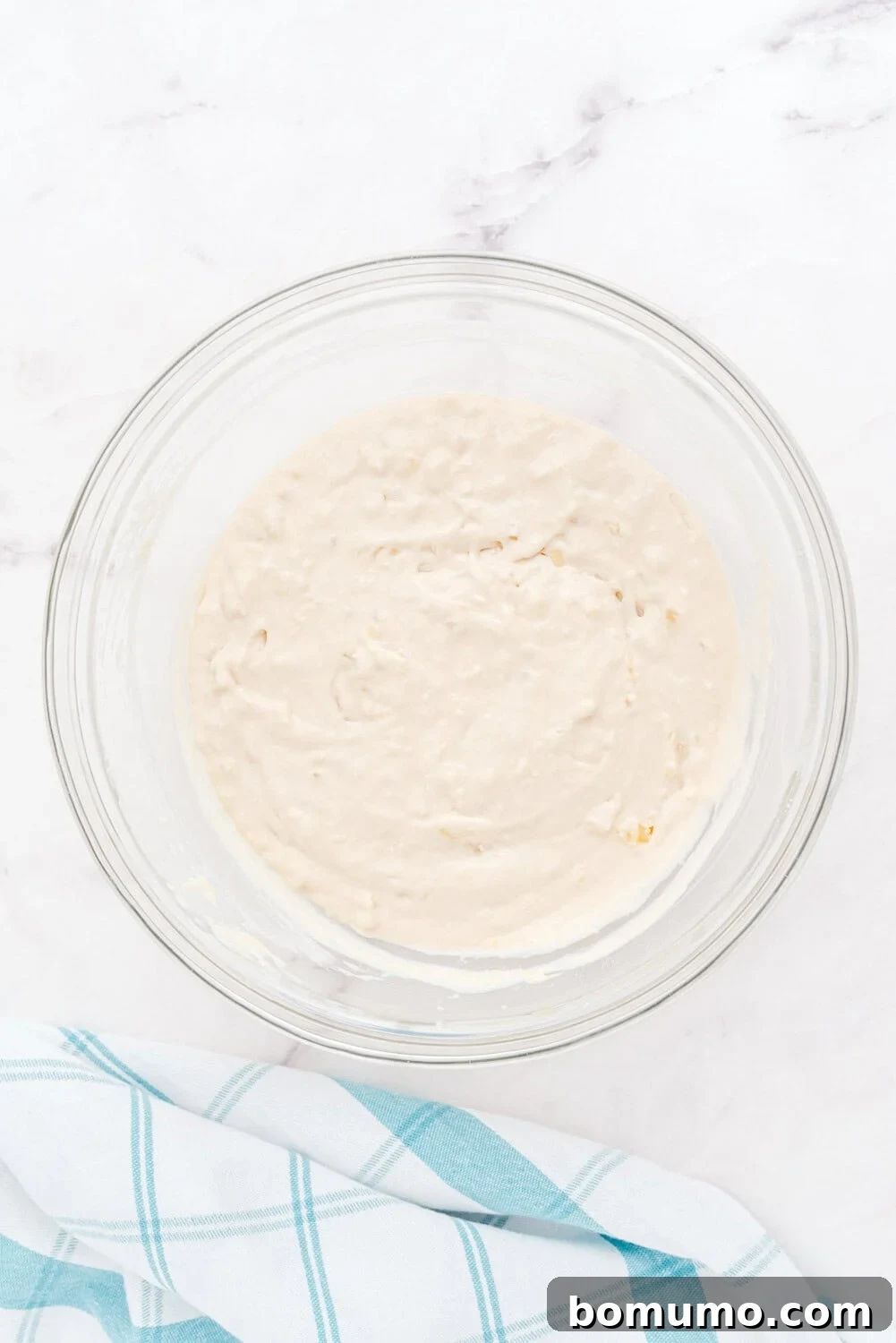 A glass bowl brimming with thick, creamy, light beige pancake batter, adorned with a generous layer of finely chopped pineapple on top, showcasing the key tropical ingredient. It rests on a spotless white surface next to a charming blue and white checkered cloth.