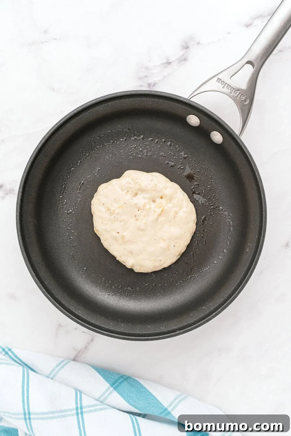 A portion of raw pancake batter gently bubbling and cooking in a nonstick frying pan, positioned on a clean white marble surface. A blue and white striped cloth is nearby, adding a pop of color to the kitchen scene.