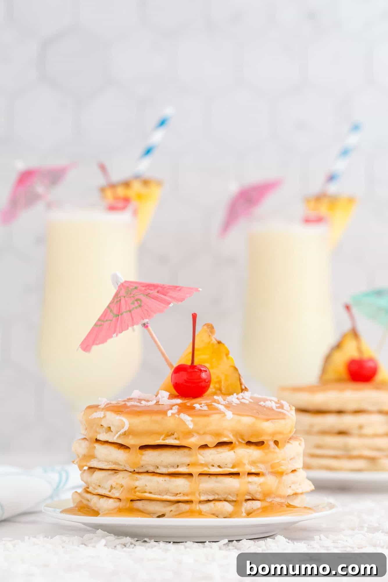 A magnificent stack of golden pancakes, lavishly topped with shimmering syrup, delicate shredded coconut, a vibrant pineapple slice, a bright red cherry, and a whimsical paper drink umbrella. In the blurred background, two frosty piña colada drinks complete the irresistible tropical scene.