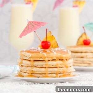 A stack of golden brown piña colada pancakes, topped with a rich caramel-like pineapple rum sauce, a vibrant pineapple slice, a bright cherry, and delicate shredded coconut, with festive paper drink umbrellas in the background.