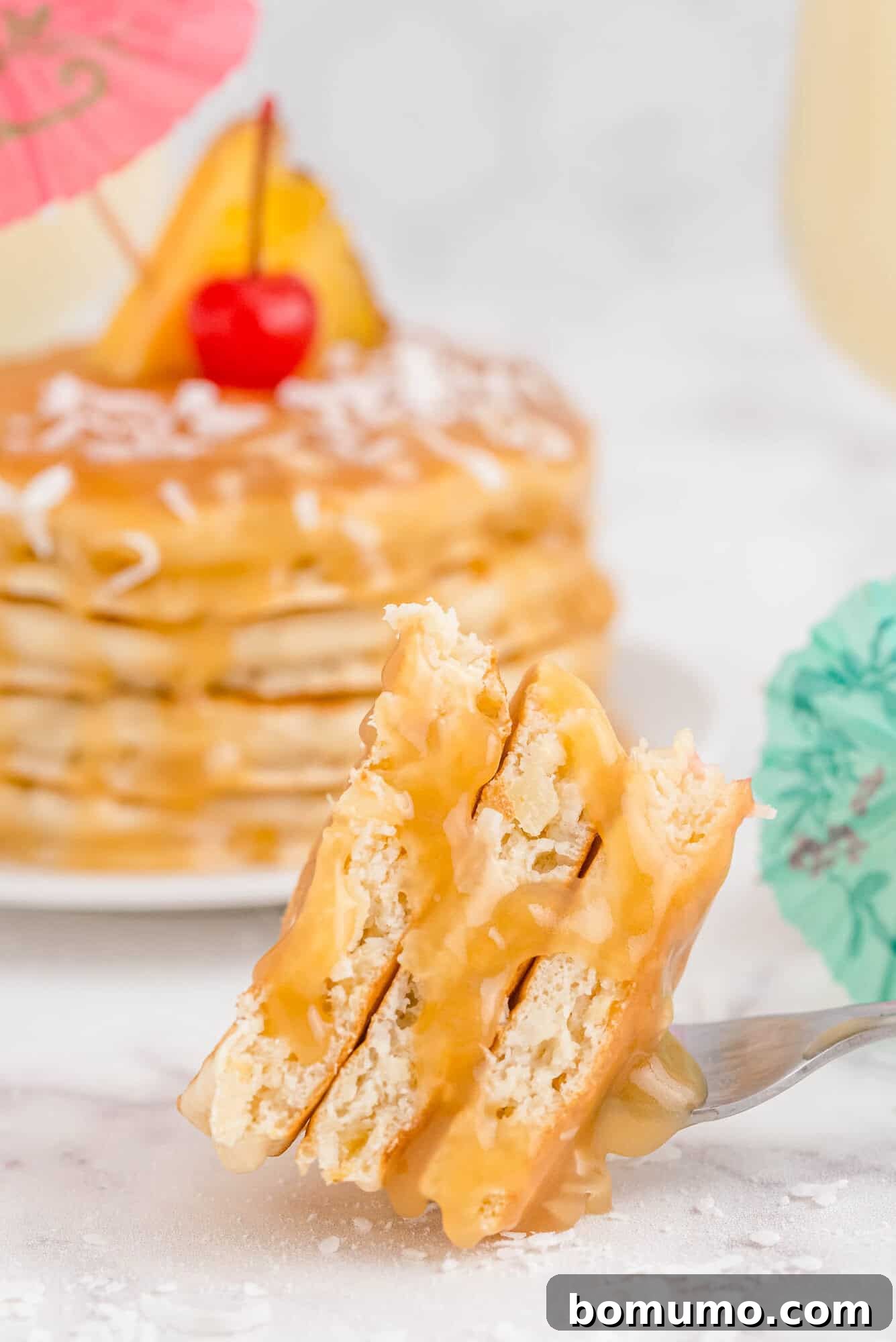 A fork lifts a golden bite of pancake, glistening with warm pineapple rum sauce. In the soft-focus background, a towering stack of beautifully decorated piña colada pancakes awaits, adorned with fresh fruit and miniature cocktail umbrellas, completing the tropical fantasy.