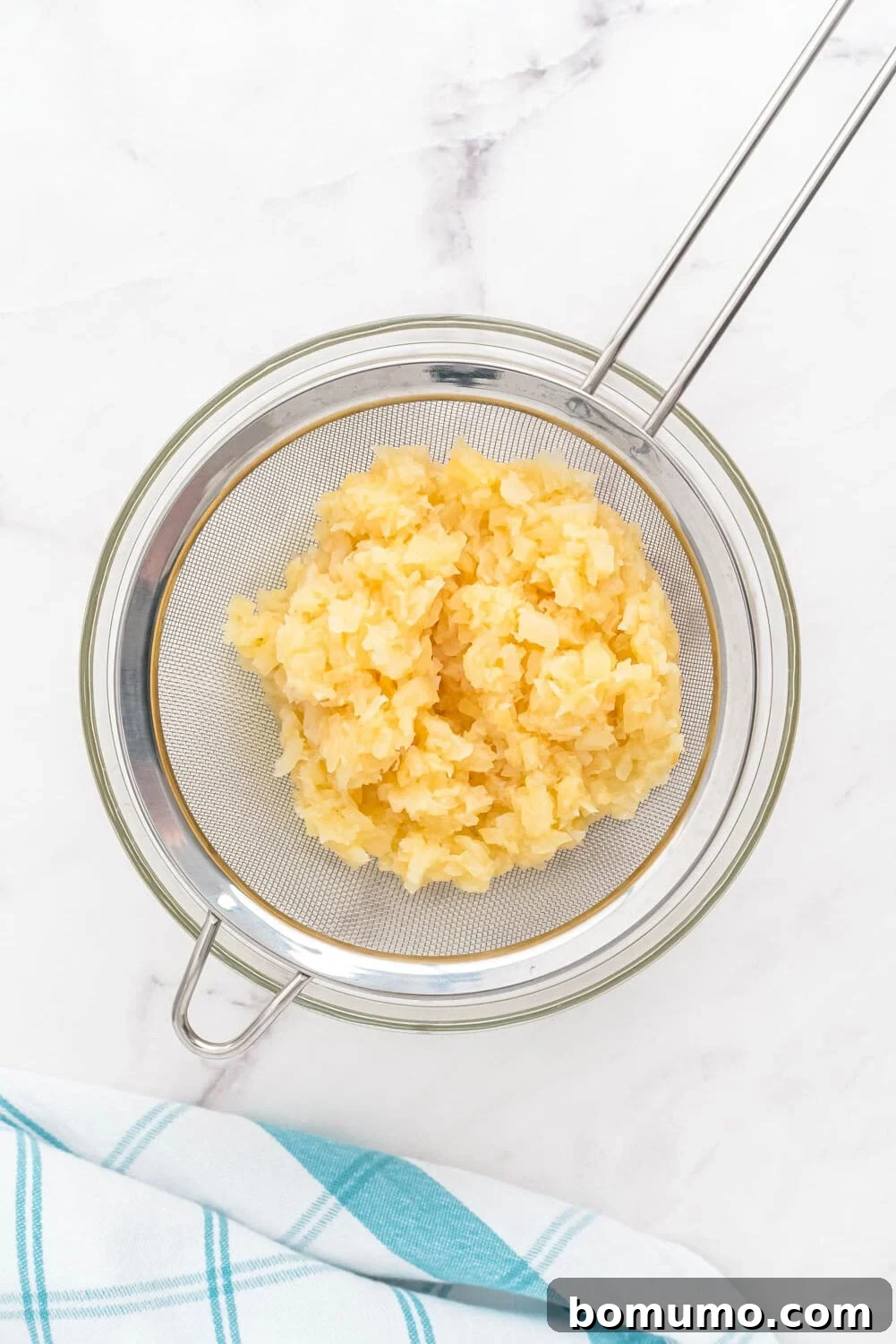 A metal strainer with thoroughly drained crushed pineapple rests over a clear glass bowl, collecting the rich juice. The setup is on a clean white surface with a neatly folded blue and white striped cloth nearby, illustrating the careful preparation process.