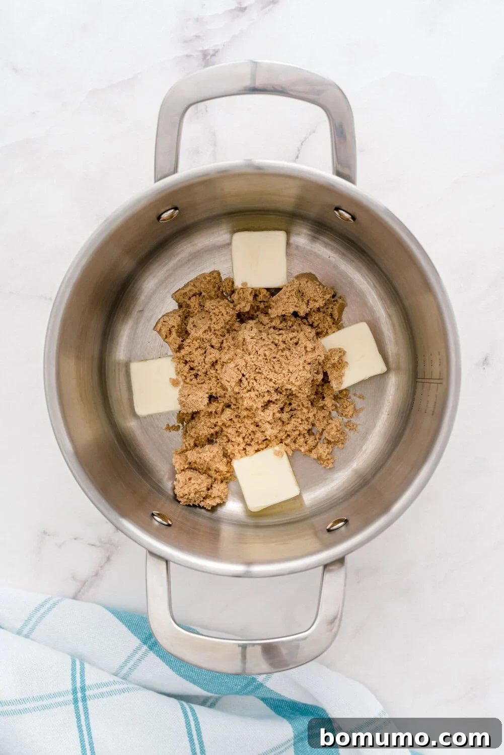 A glistening stainless steel saucepan on a bright white surface, holding a melting mixture of five pats of unsalted butter and a mound of rich brown sugar, beginning to transform into the base of a luscious sauce. A small section of a blue-striped kitchen cloth is visible, adding a homely touch.