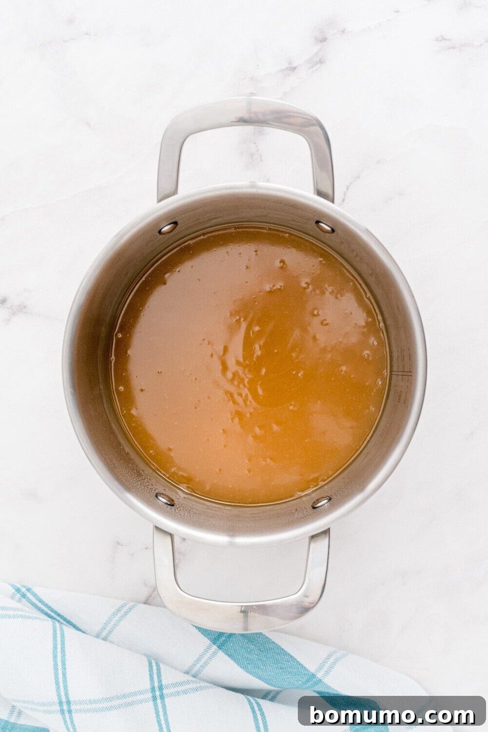 A stainless steel pot filled with a rich, bubbling brown liquid — the pineapple rum sauce — sits on a pristine white surface, indicating it's nearly ready. A blue and white checkered kitchen towel provides a subtle accent in the background.