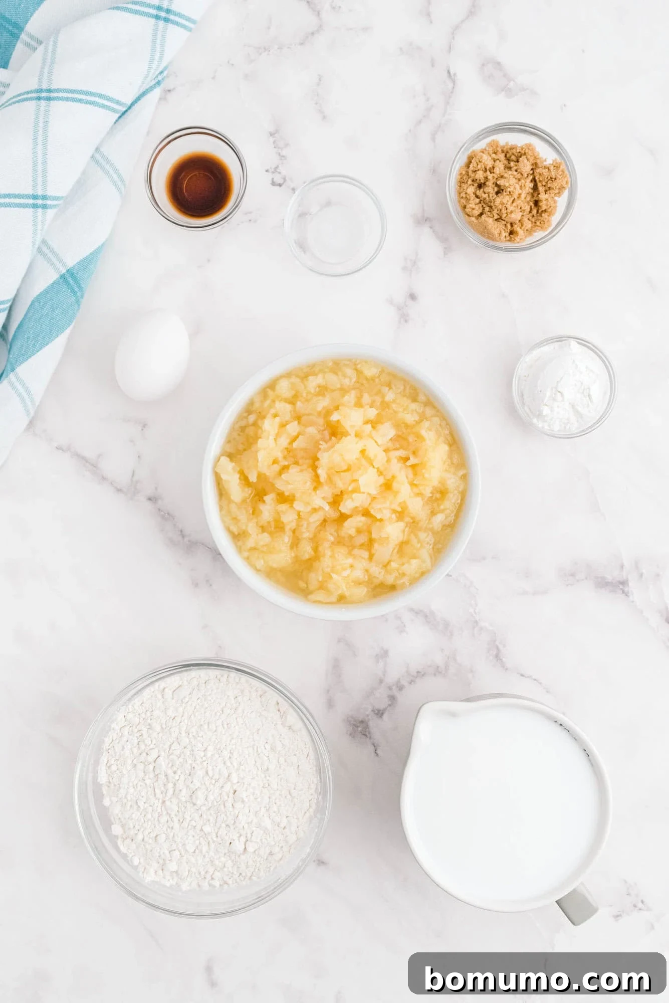 A beautifully arranged selection of fresh ingredients for Piña Colada Pancakes on a white marble surface, including fluffy flour, creamy coconut milk, rich brown sugar, a single egg, vanilla extract, a bowl of crushed pineapple, baking powder, and salt. A blue and white striped towel sits neatly to the side, highlighting the meticulous preparation.