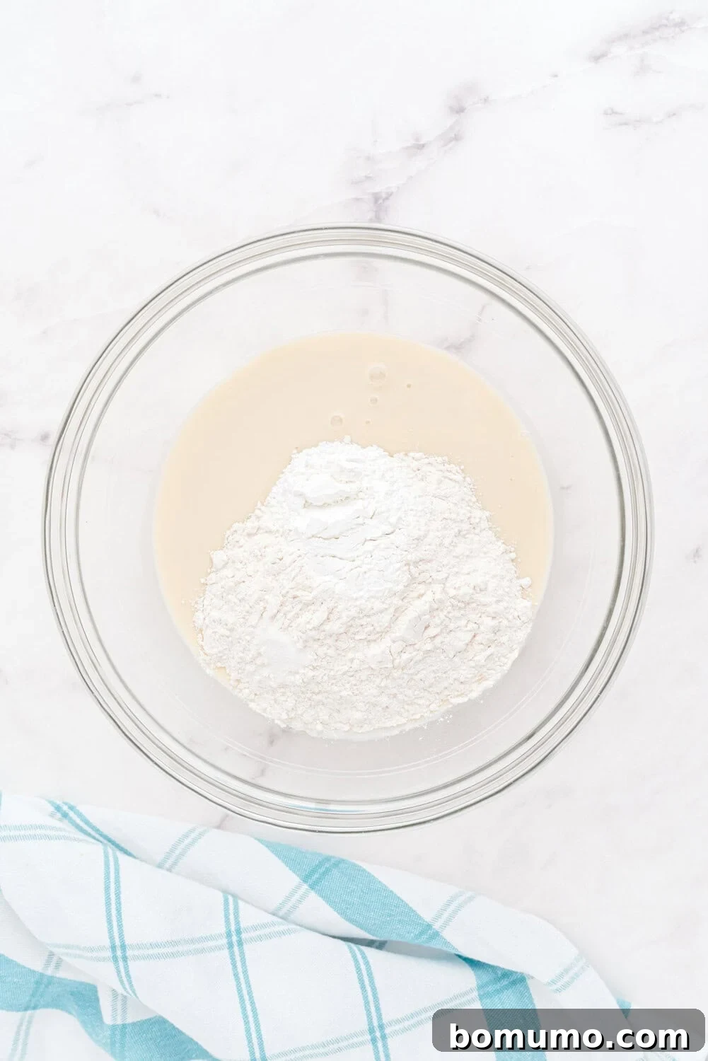 A transparent glass bowl on a pristine white marble surface, holding a creamy mixture of coconut milk and flour, hinting at the start of a delightful pancake batter. A blue and white checkered kitchen towel is placed nearby, adding a touch of charm.