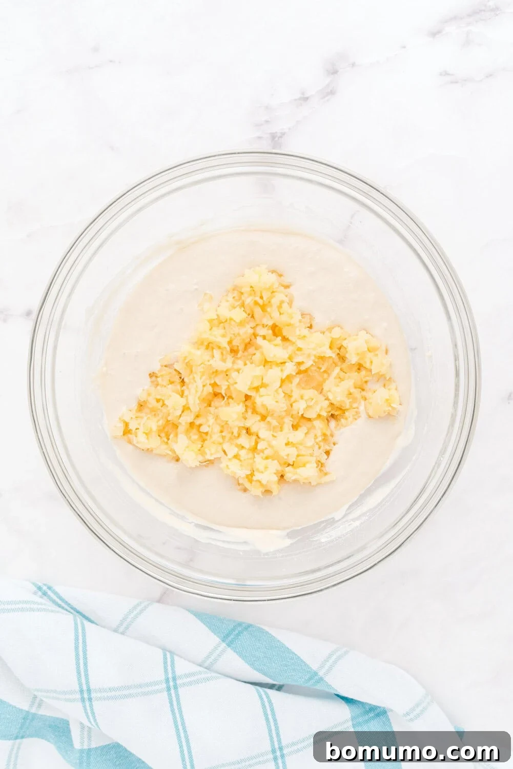 A glass bowl filled with rich, light beige pancake batter. The texture is thick and inviting, promising perfectly fluffy results. A blue and white checkered kitchen towel rests next to the bowl on a pristine white surface.