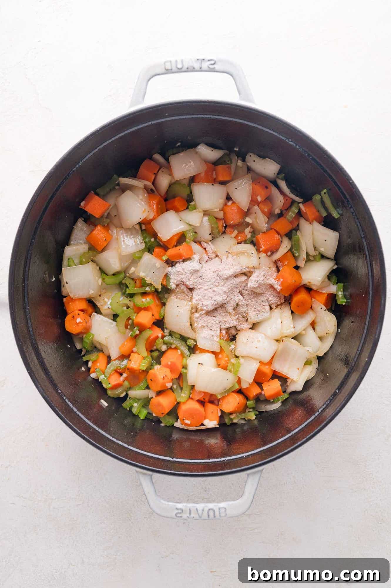 Tender Pork Ragout 11 Chopped onions, carrots, and celery sautéing in a black pot, with a mound of seasoned flour added on top, ready to be incorporated.