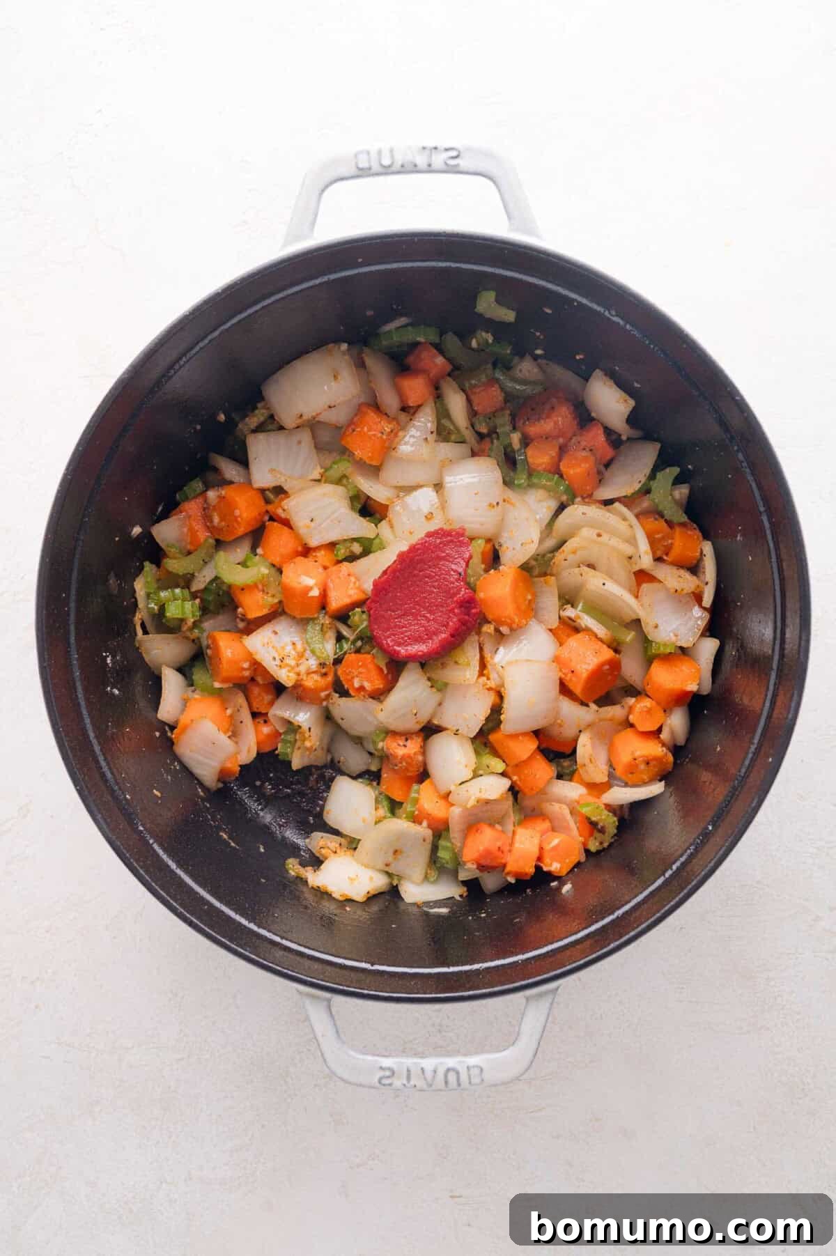 Tender Pork Ragout 13 Chopped onions, carrots, and celery with a dollop of vibrant tomato paste in a black pot, viewed from above, ready to be stirred in.