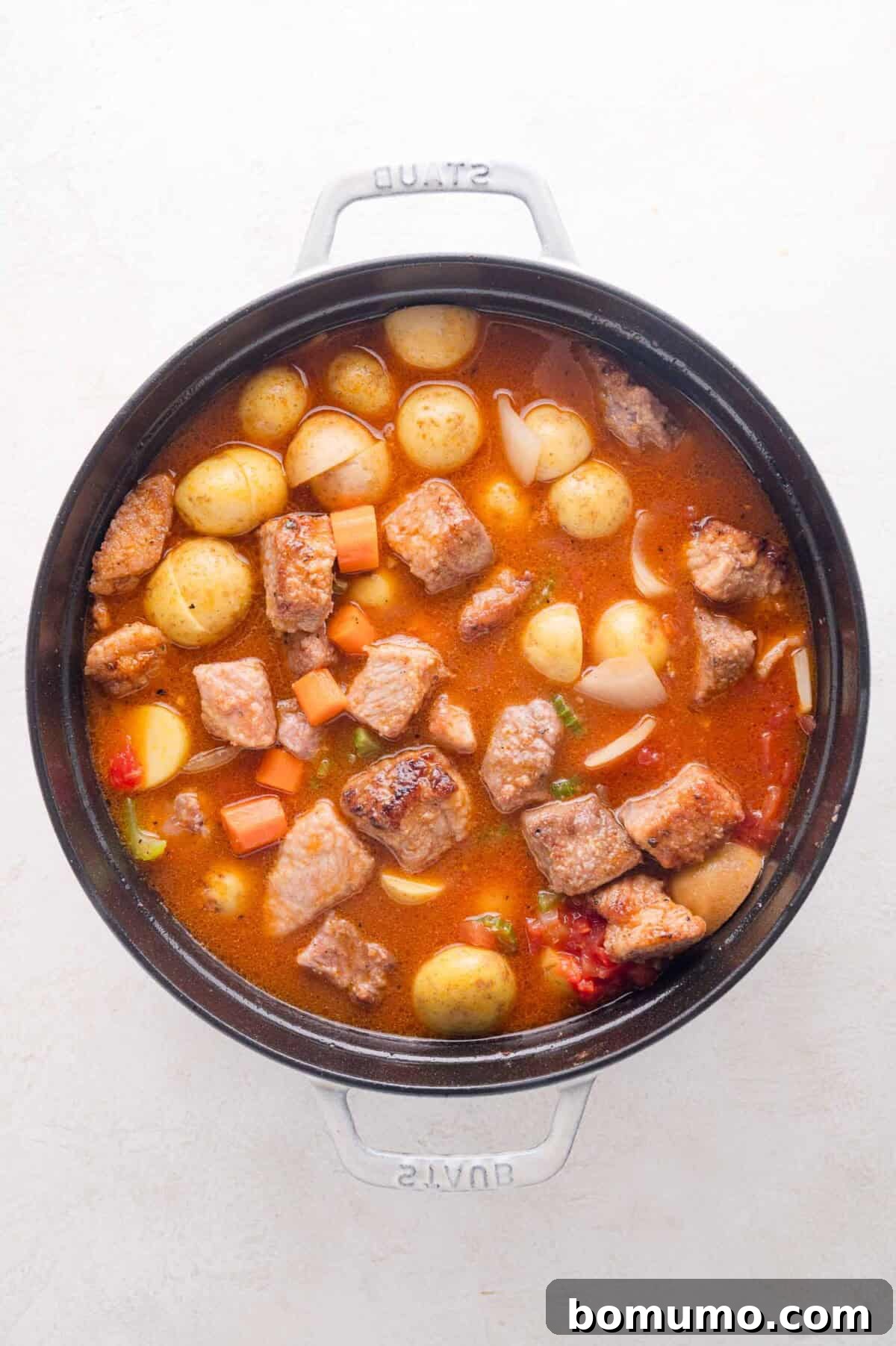 Tender Pork Ragout 14 A pot of rich pork stew with succulent chunks of pork, whole baby potatoes, carrots, onions, and broth, viewed from above on a white background, showcasing ingredients before simmering.
