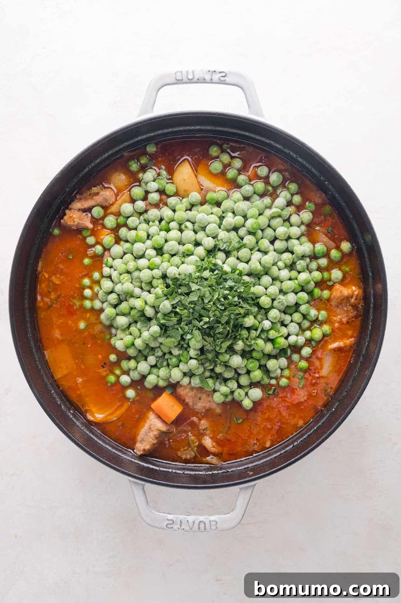 Tender Pork Ragout 17 A pot of savory stew containing pork, potatoes, carrots, and a rich tomato sauce, topped with a pile of frozen green peas and freshly chopped parsley, ready for the final simmer.