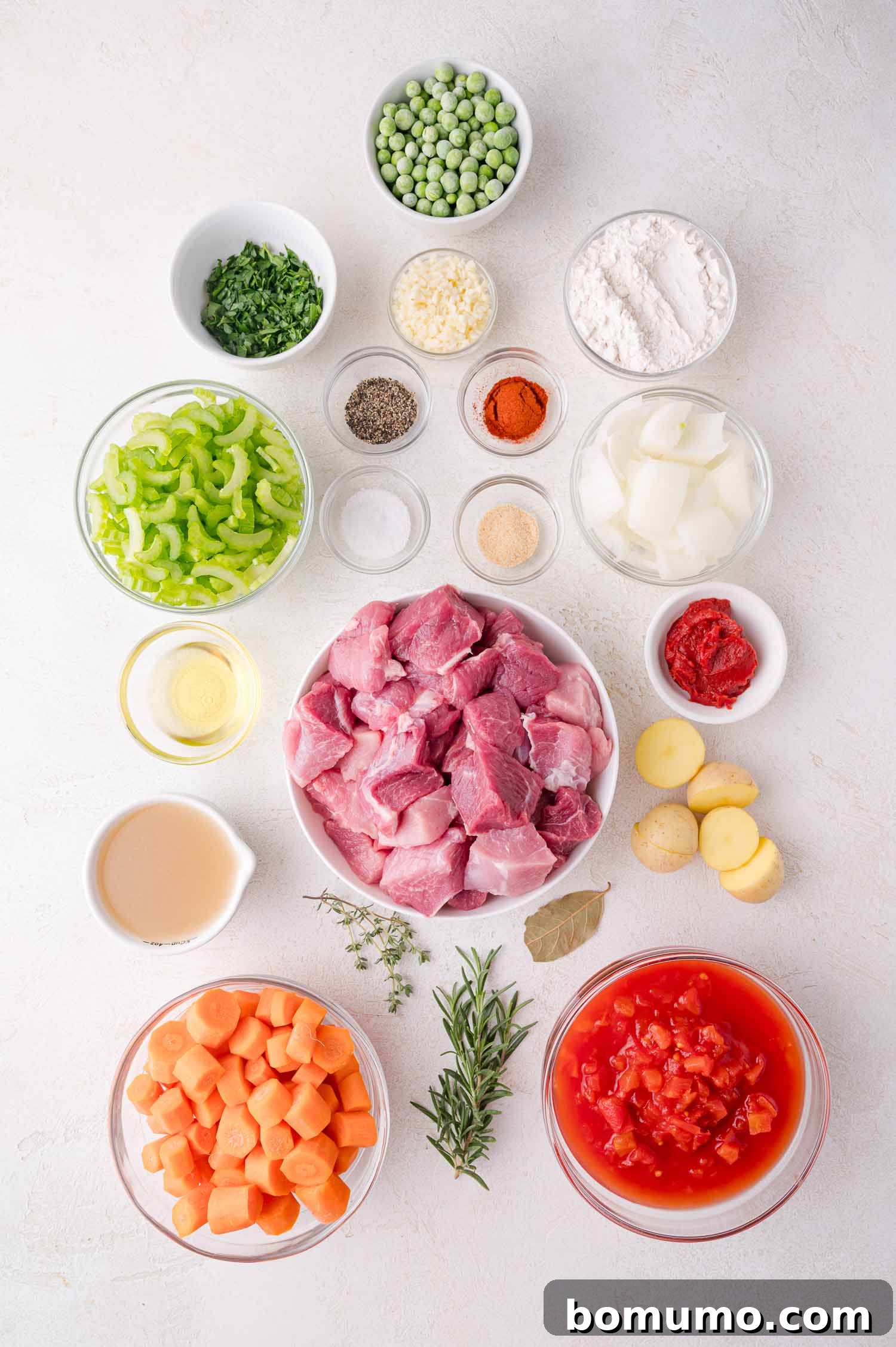 Tender Pork Ragout 4 Assorted stew ingredients including raw pork, diced tomatoes, carrots, celery, peas, potatoes, onions, spices, herbs, tomato paste, oil, and flour arranged in bowls on a white surface, ready for cooking.