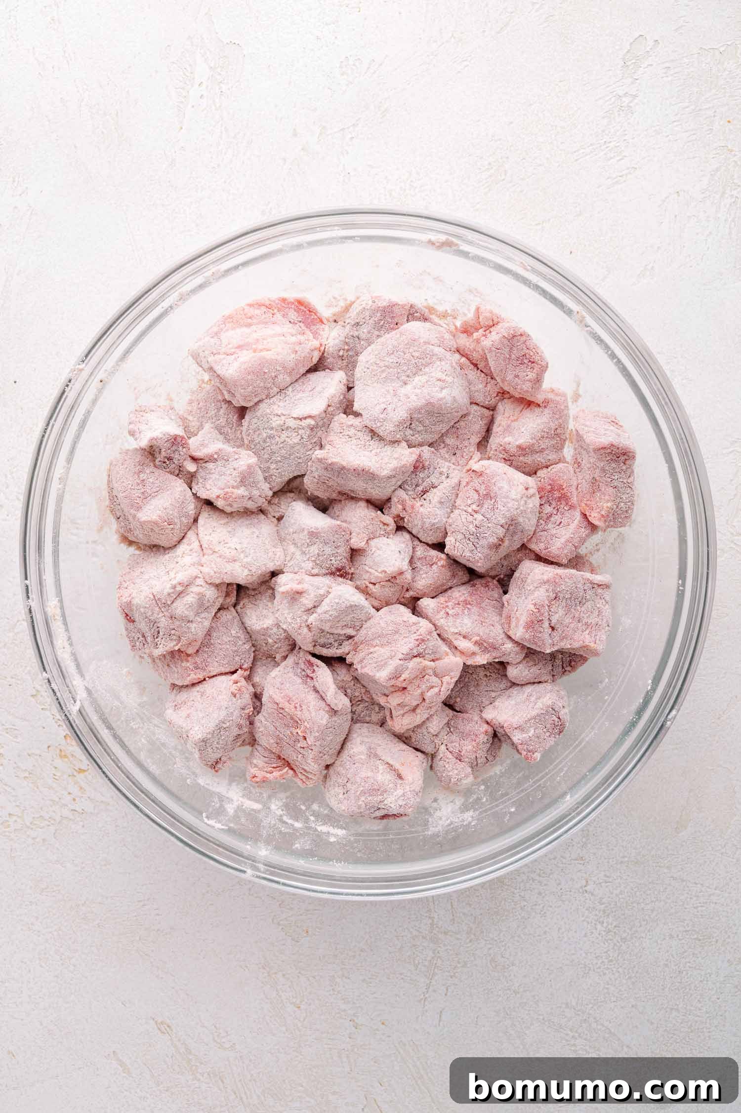 Tender Pork Ragout 6 A glass bowl filled with raw pork cubes generously coated in flour, ready for searing, presented on a light-colored surface.