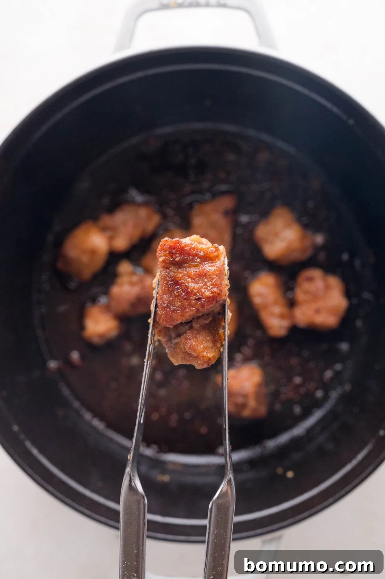 Tender Pork Ragout 8 A pair of metal tongs holding a piece of cooked, browned pork above a black pot with more pieces of meat inside, illustrating the searing process.