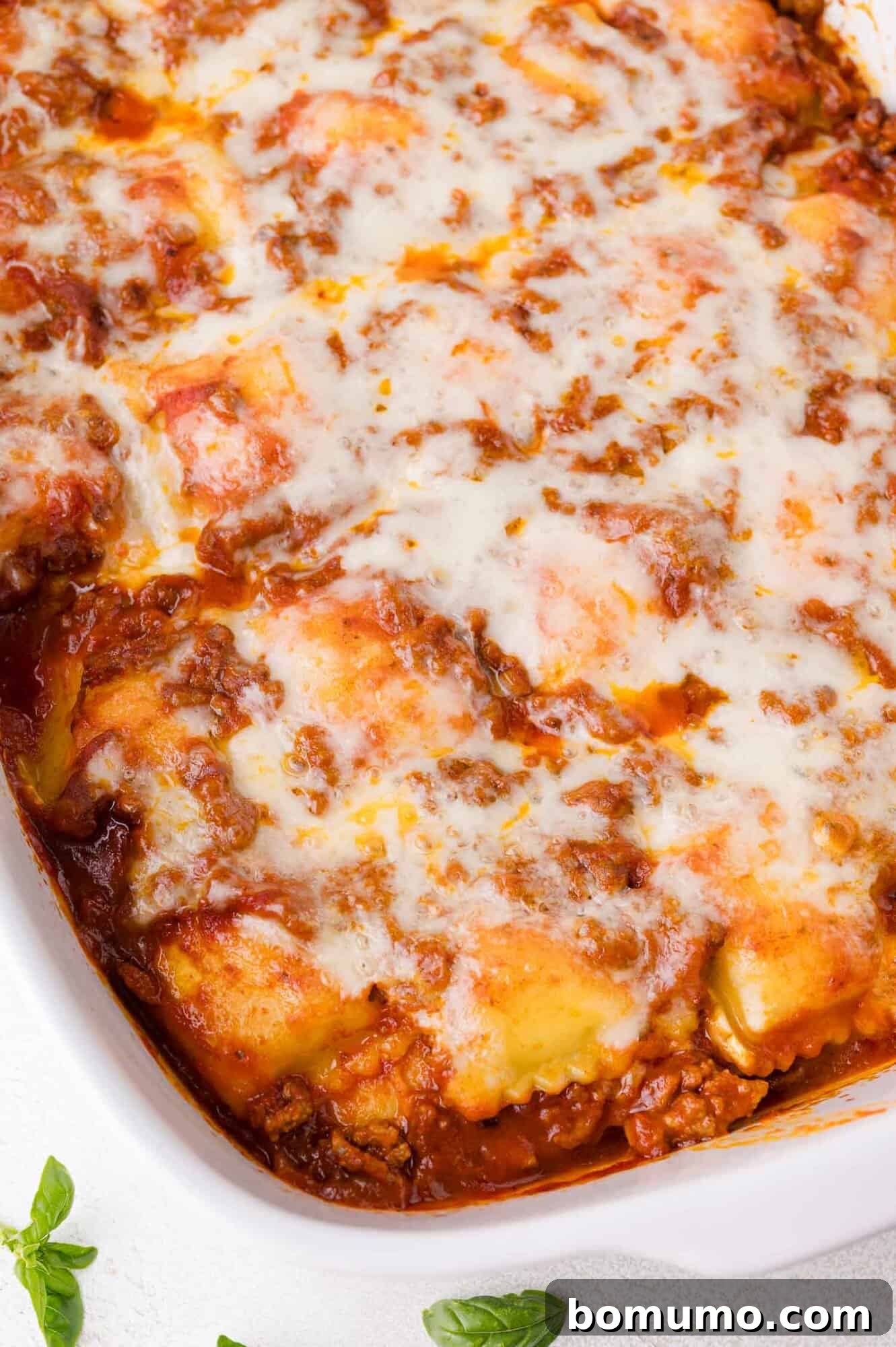 A vibrant close-up of baked ravioli, golden-brown and bubbling in a white casserole dish. Layers of rich meat sauce and tender pasta are visible beneath a generous topping of perfectly melted, gooey cheese, creating an irresistible texture and aroma.