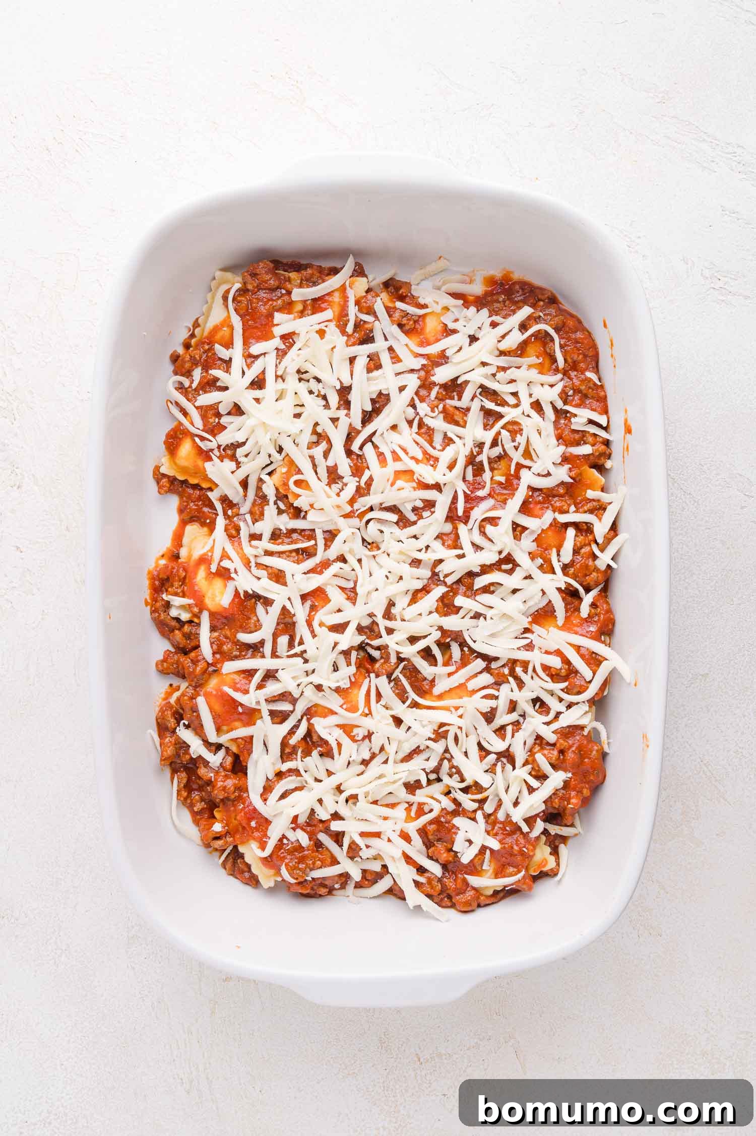 A pristine white baking dish, viewed from directly above, is filled with layers of uncooked lasagna, lush shredded cheese, and rich tomato meat sauce, meticulously arranged and ready for baking.