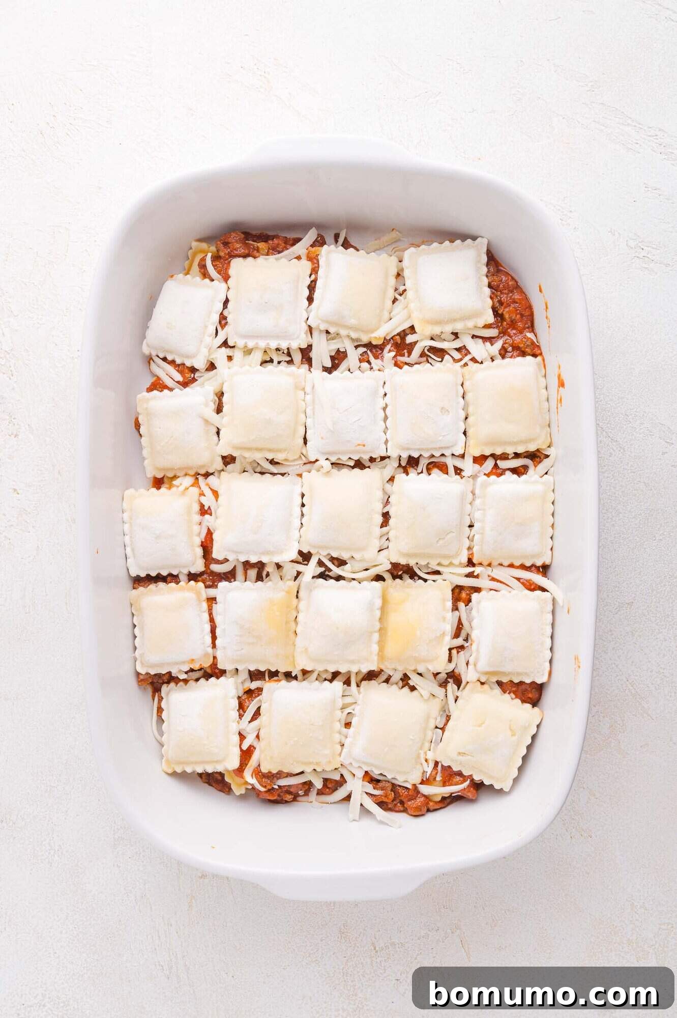 A pristine white baking dish holds an inviting combination of ground meat sauce, a generous layer of shredded cheese, and uncooked square ravioli, all arranged in neat rows and ready to be transformed in the oven.
