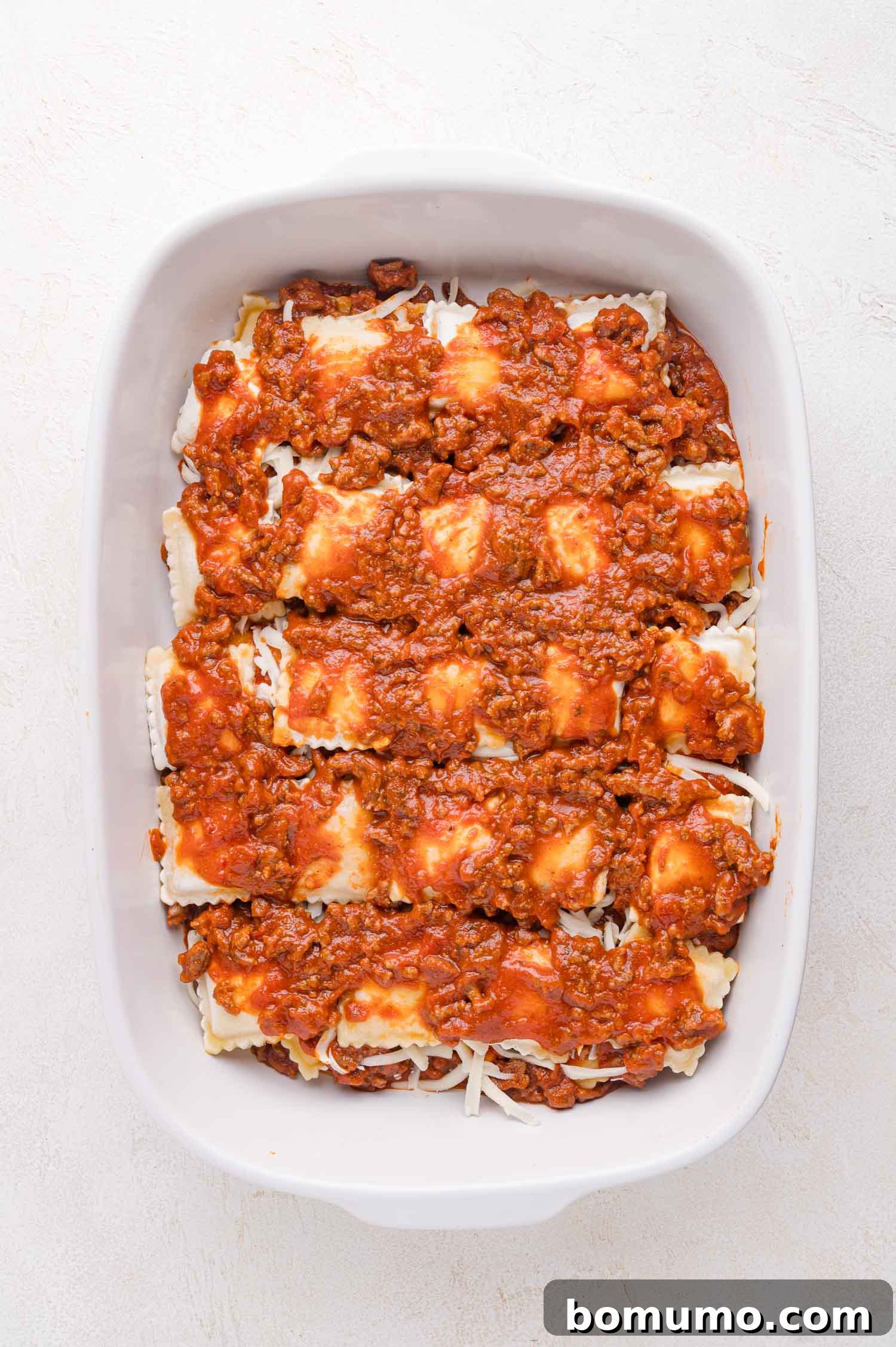 A white baking dish is meticulously filled with enticing layers of ravioli, shredded cheese, and rich meat sauce, perfectly prepped and awaiting its delicious transformation in the oven.