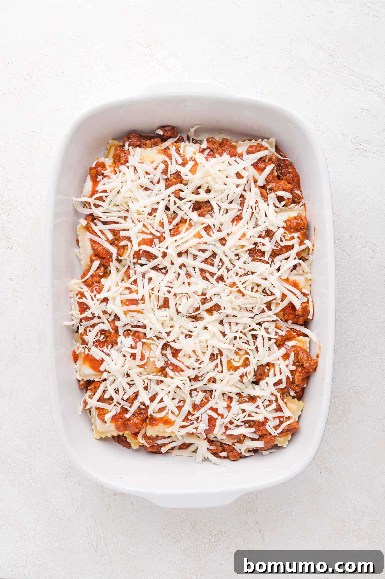 A pristine white baking dish is filled with perfectly layered baked ravioli, rich meat sauce, and glistening shredded cheese, beautifully assembled and ready for the oven to create a comforting meal.