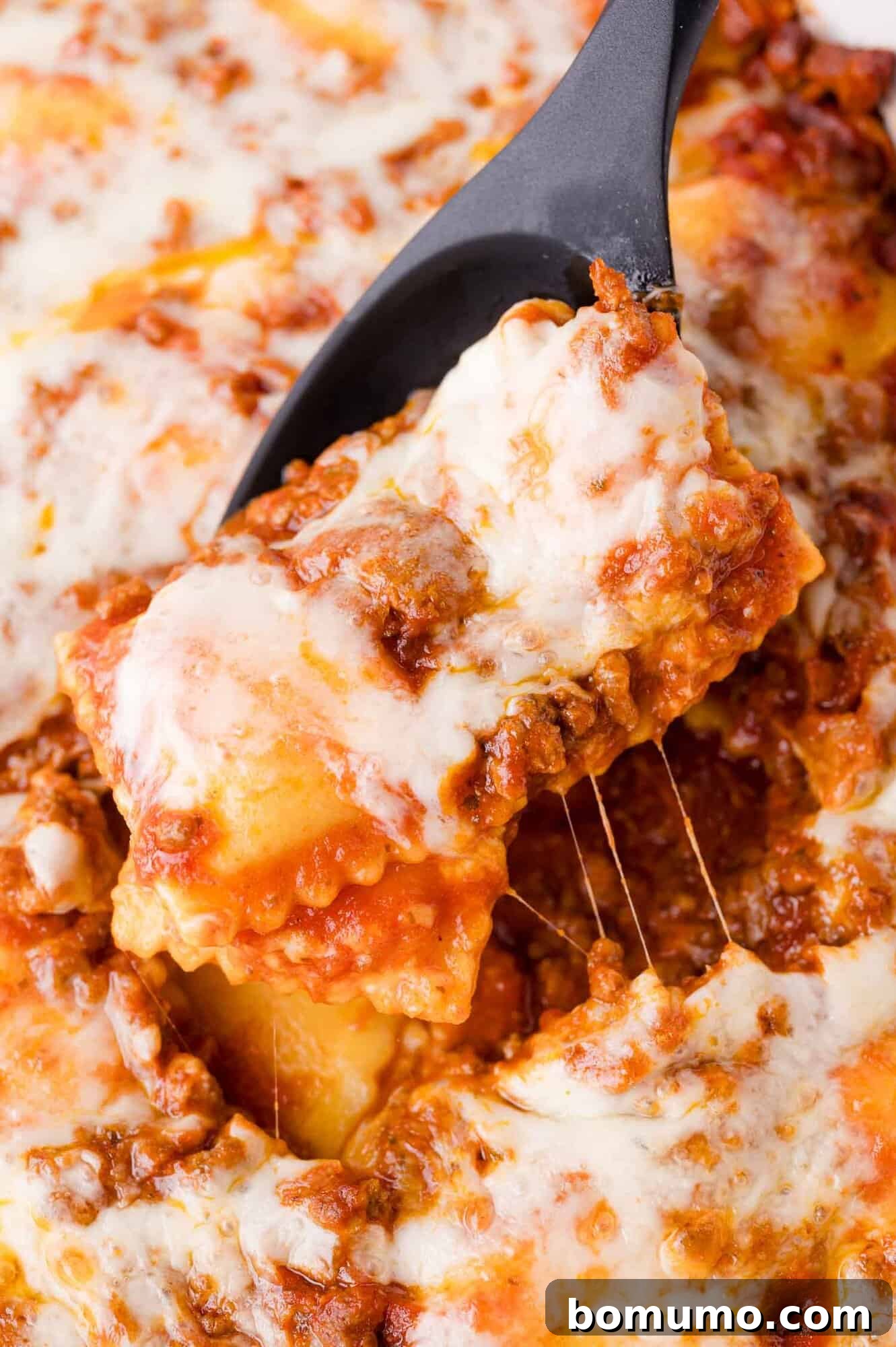 A close-up shot captures a serving spoon expertly lifting a generous portion of cheesy, meat-filled ravioli from a baked pasta dish. The spoon reveals abundant melted cheese, rich tomato sauce, and tender pasta, creating an irresistible visual.