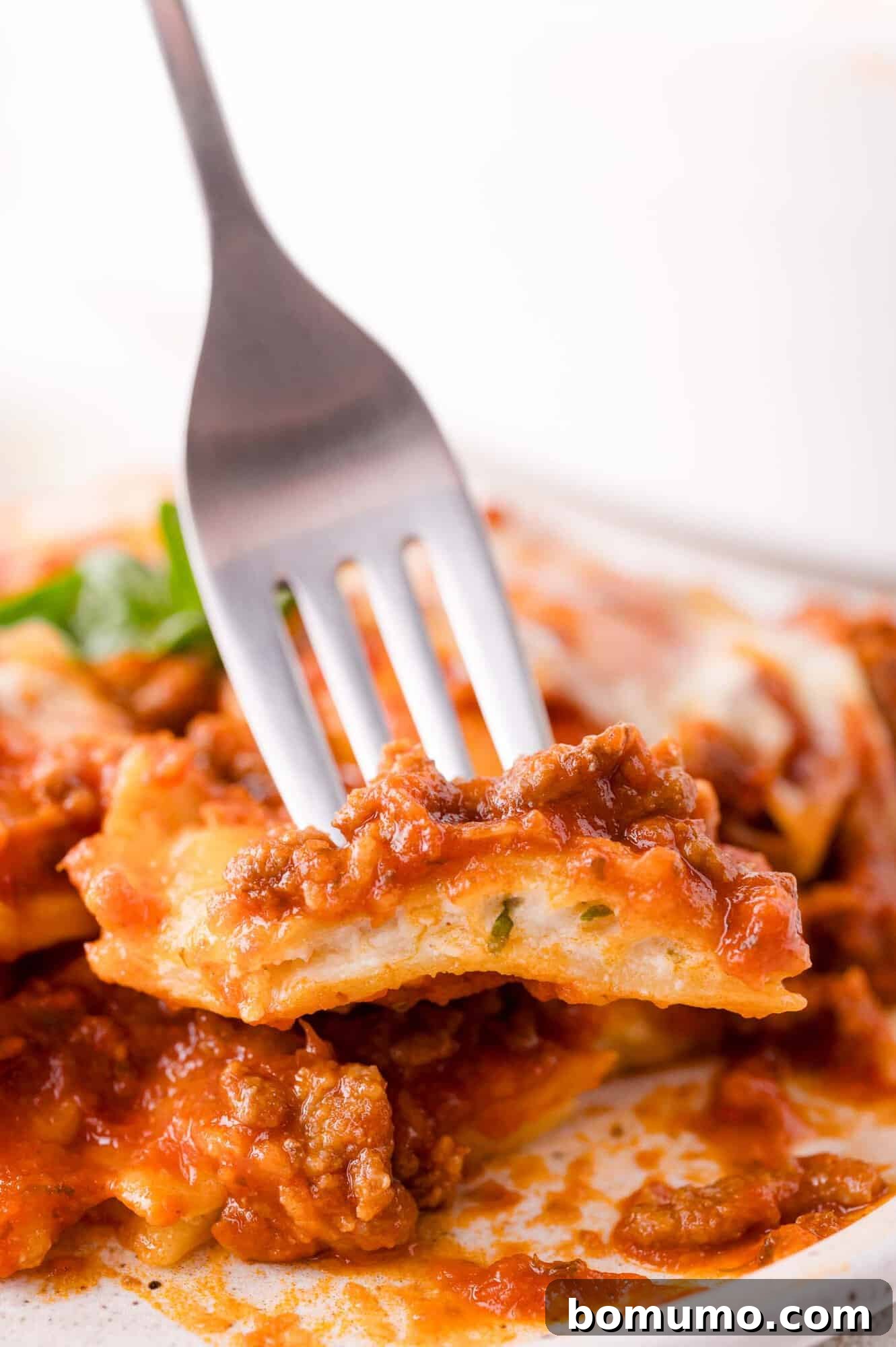 A tantalizing close-up shows a fork holding a single piece of baked ravioli, revealing luscious meat sauce, melted cheese, and a sprinkle of fresh parsley, perfectly presented on a white plate.