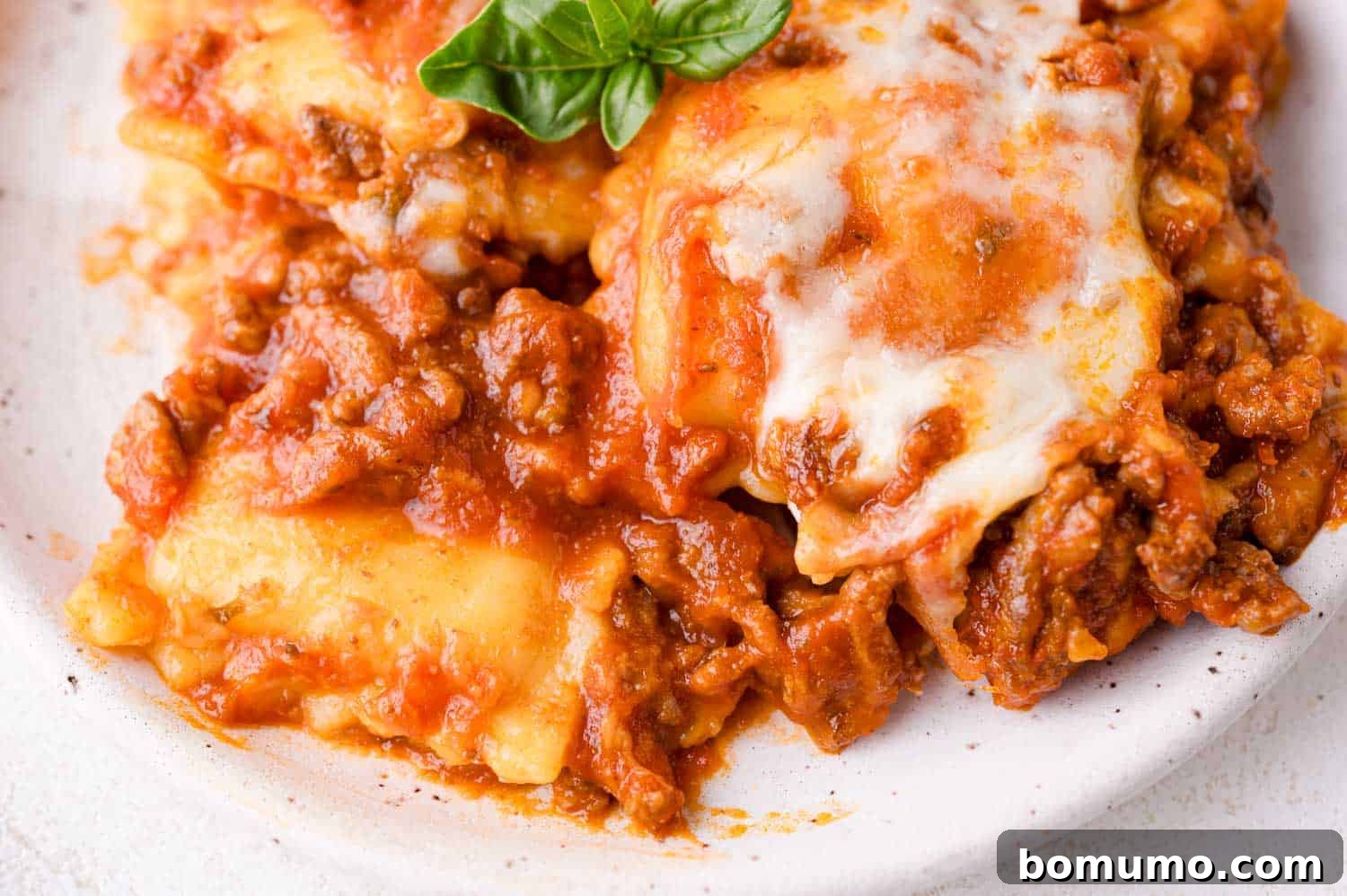 A mouth-watering close-up of a perfectly portioned serving of baked ravioli on a pristine white plate. The dish is generously topped with a rich, melted cheese layer and a hearty tomato meat sauce, beautifully garnished with a vibrant, fresh basil leaf for color and aroma.