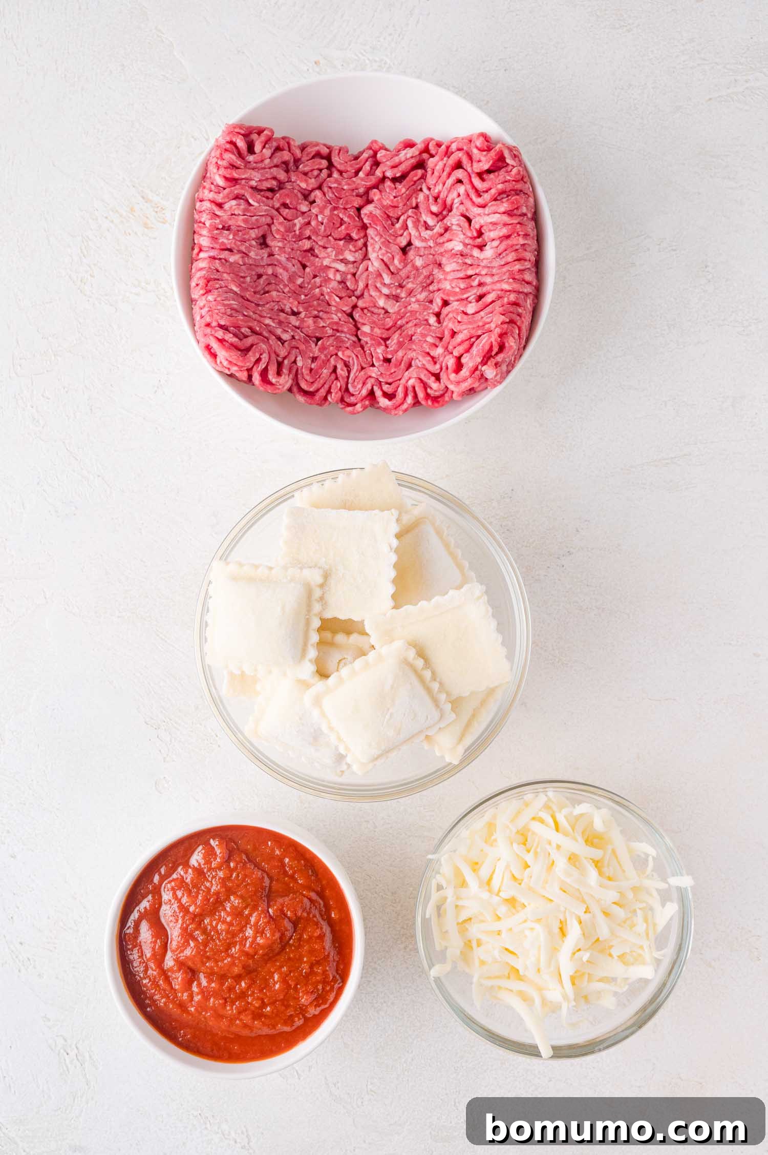 A flat lay of four bowls showcasing the simple ingredients for baked ravioli: perfectly crumbled ground beef, neatly arranged square ravioli, a rich, vibrant tomato sauce, and a pile of freshly shredded mozzarella cheese, all ready for assembly on a clean white surface.