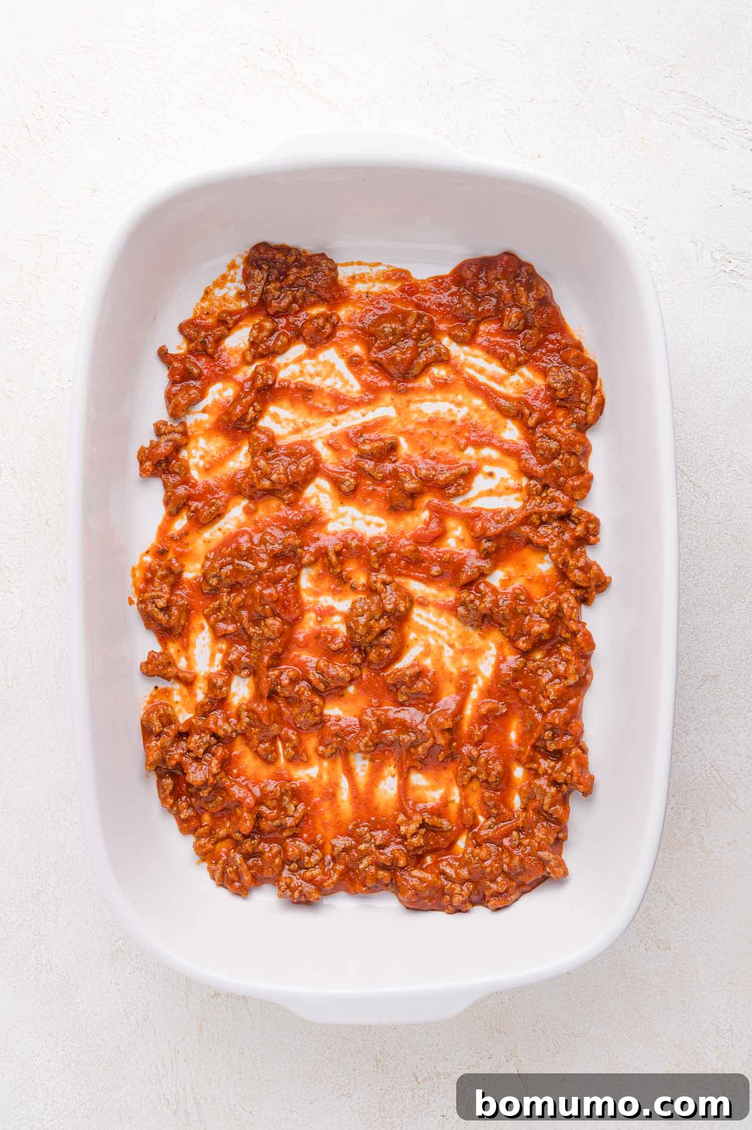 A pristine white casserole dish showcases a vibrant layer of rich, cooked ground beef mixed with tomato sauce, spread perfectly across its bottom, setting the stage for the baked ravioli.