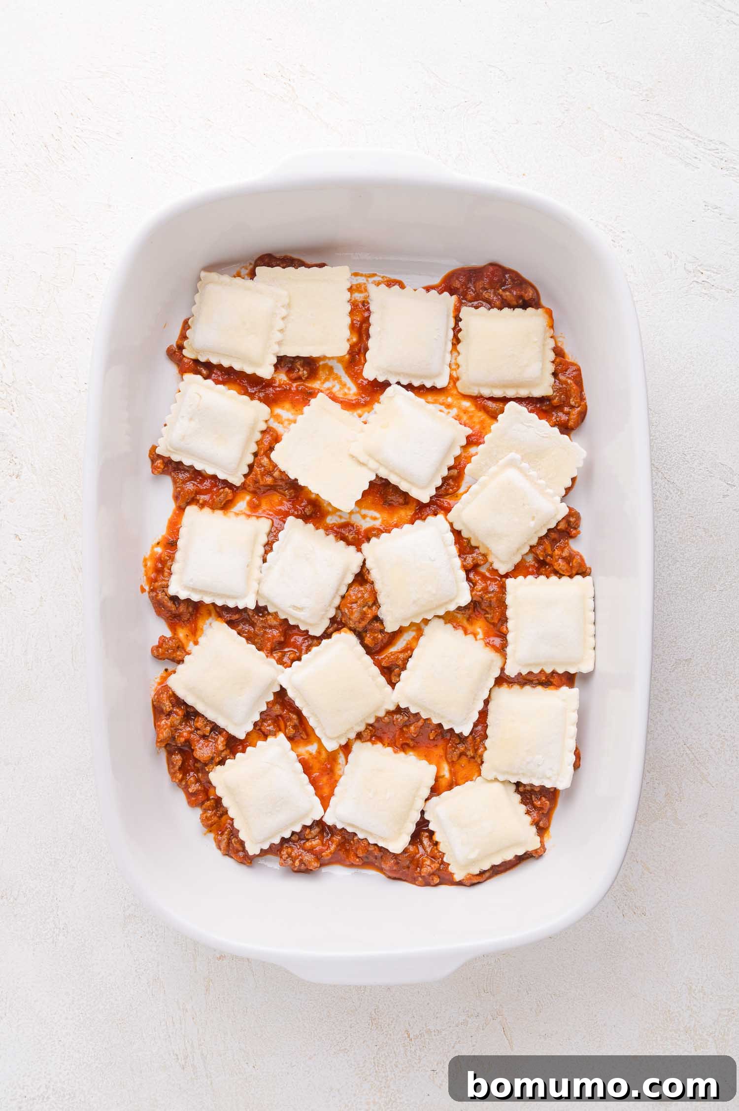 A white baking dish displays an even layer of savory meat sauce, meticulously topped with rows of uncooked ravioli, precisely arranged and ready for the next delicious layer.