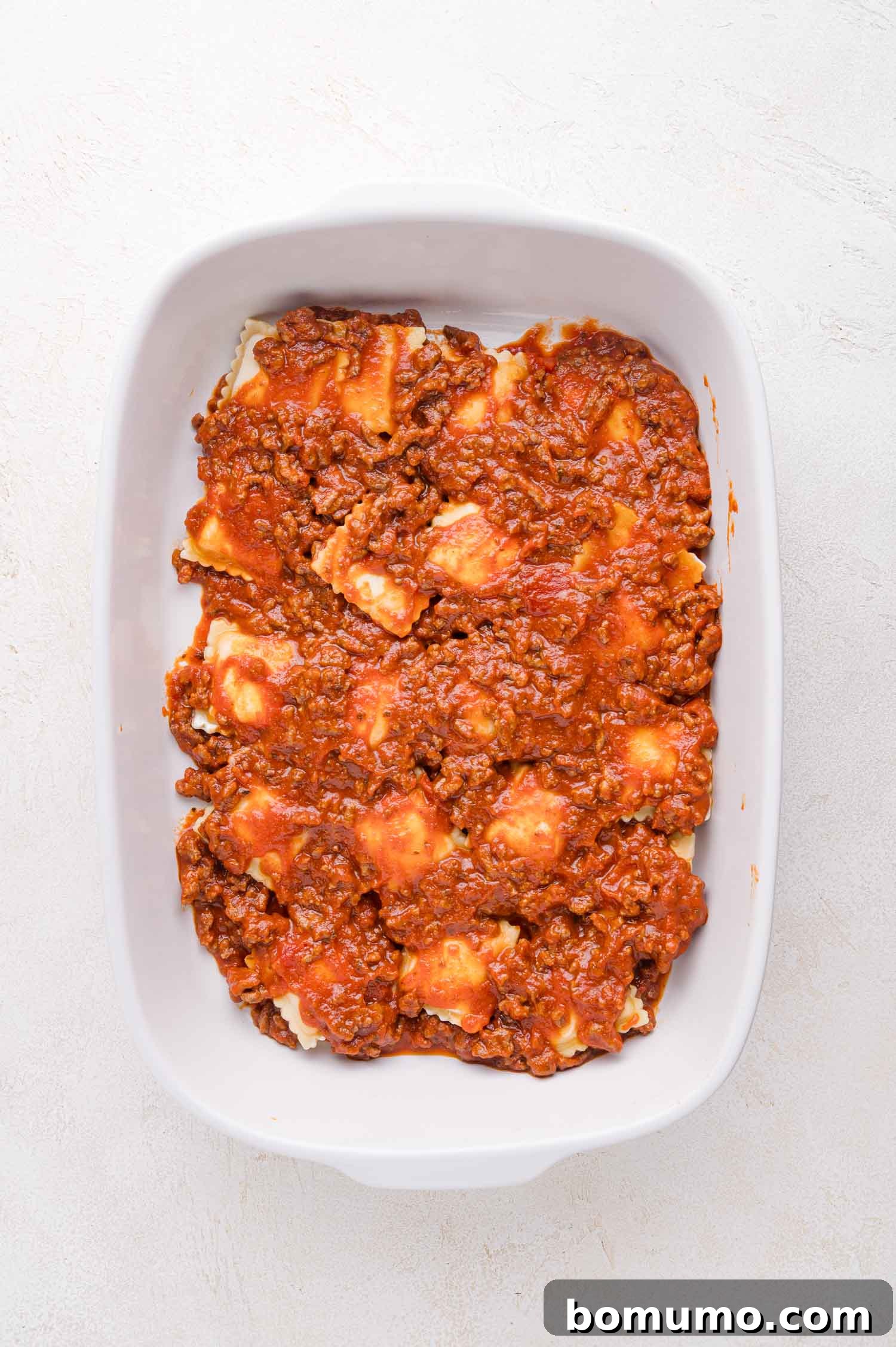 Inside a clean white baking dish, a layer of delicate ravioli is generously covered with a rich, vibrant meat sauce, creating a tantalizing visual of flavors mingling.