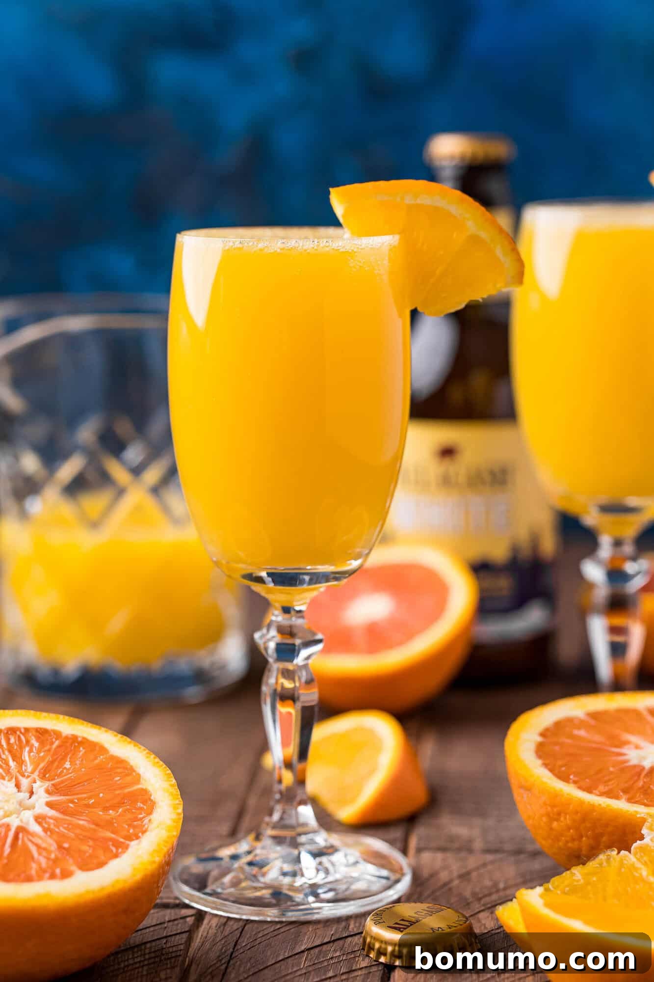 A chilled glass of beermosa, garnished with a fresh orange slice, resting on a rustic wooden table. In the background, there are artfully arranged halved and sliced oranges, alongside a bottle of wheat beer, suggesting the ingredients used for this refreshing drink.