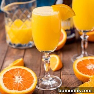 A glass of vibrant orange Beermosa, garnished with a fresh orange slice, stands on a wooden table. In the background, sliced oranges and a pitcher of juice provide context, highlighting the refreshing nature of the drink.