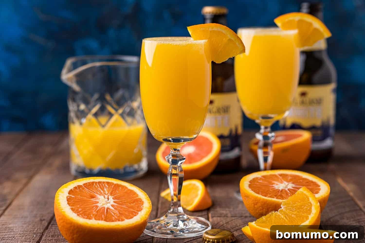 Two elegantly presented glasses of Beermosa, each adorned with a vibrant orange slice, sit on a polished wooden table. In the background, fresh halved oranges, a pitcher of orange juice, and bottles of wheat beer are visible, creating a refreshing scene ready for a brunch gathering.