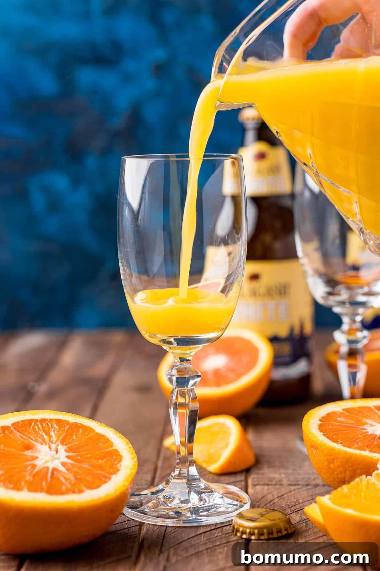 A hand carefully pours bright orange juice from a clear pitcher into a elegant wine glass. The scene is set on a wooden table, with fresh orange slices and a bottle of beer blurred in the background, illustrating the first step of making a Beermosa.