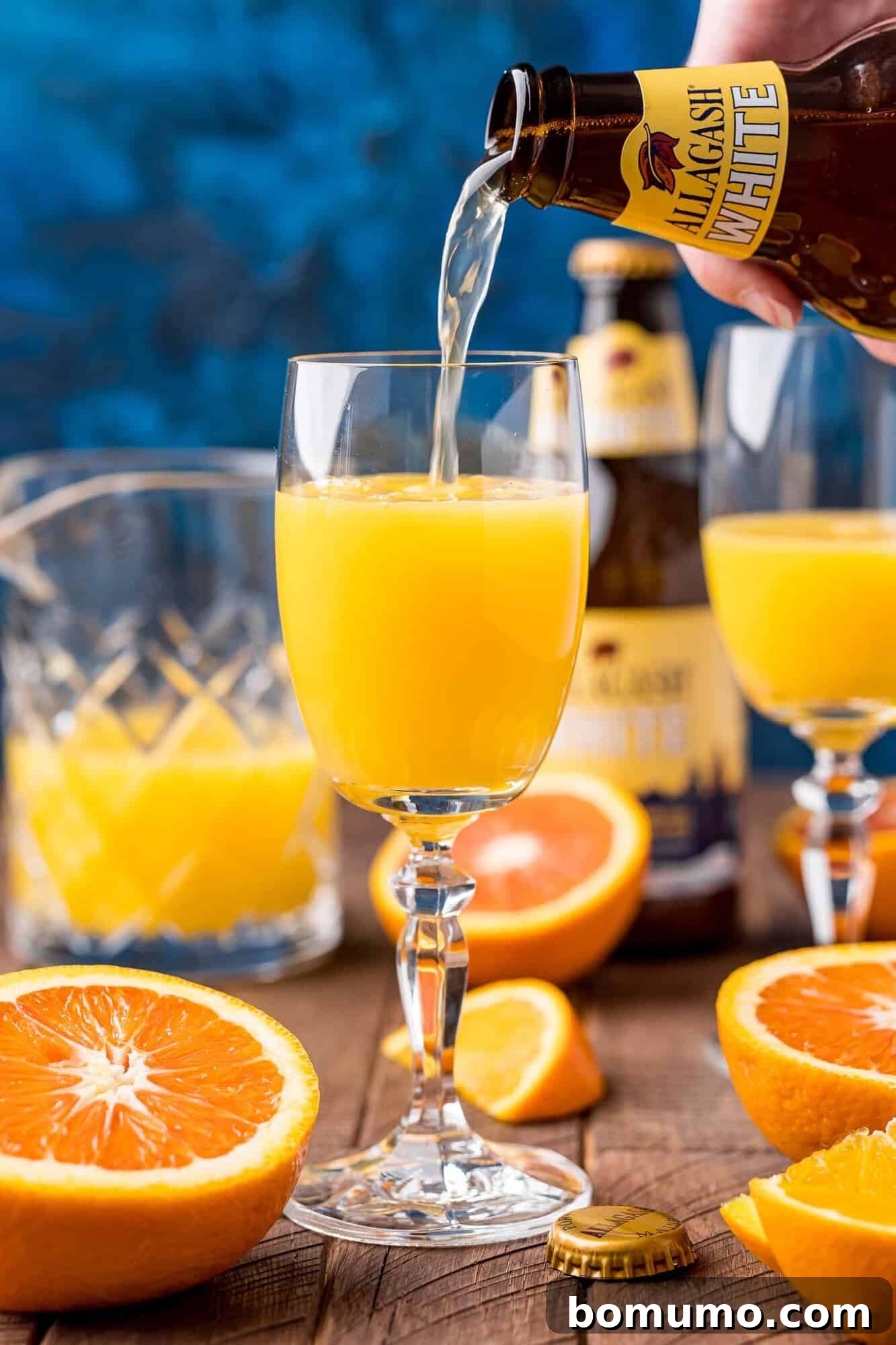 A hand gently tilts a bottle of Allagash White beer, pouring its contents into a glass already containing orange juice. The beer cascades, creating a vibrant Beermosa. On the wooden table, sliced oranges, another glass, and a second beer bottle are present.