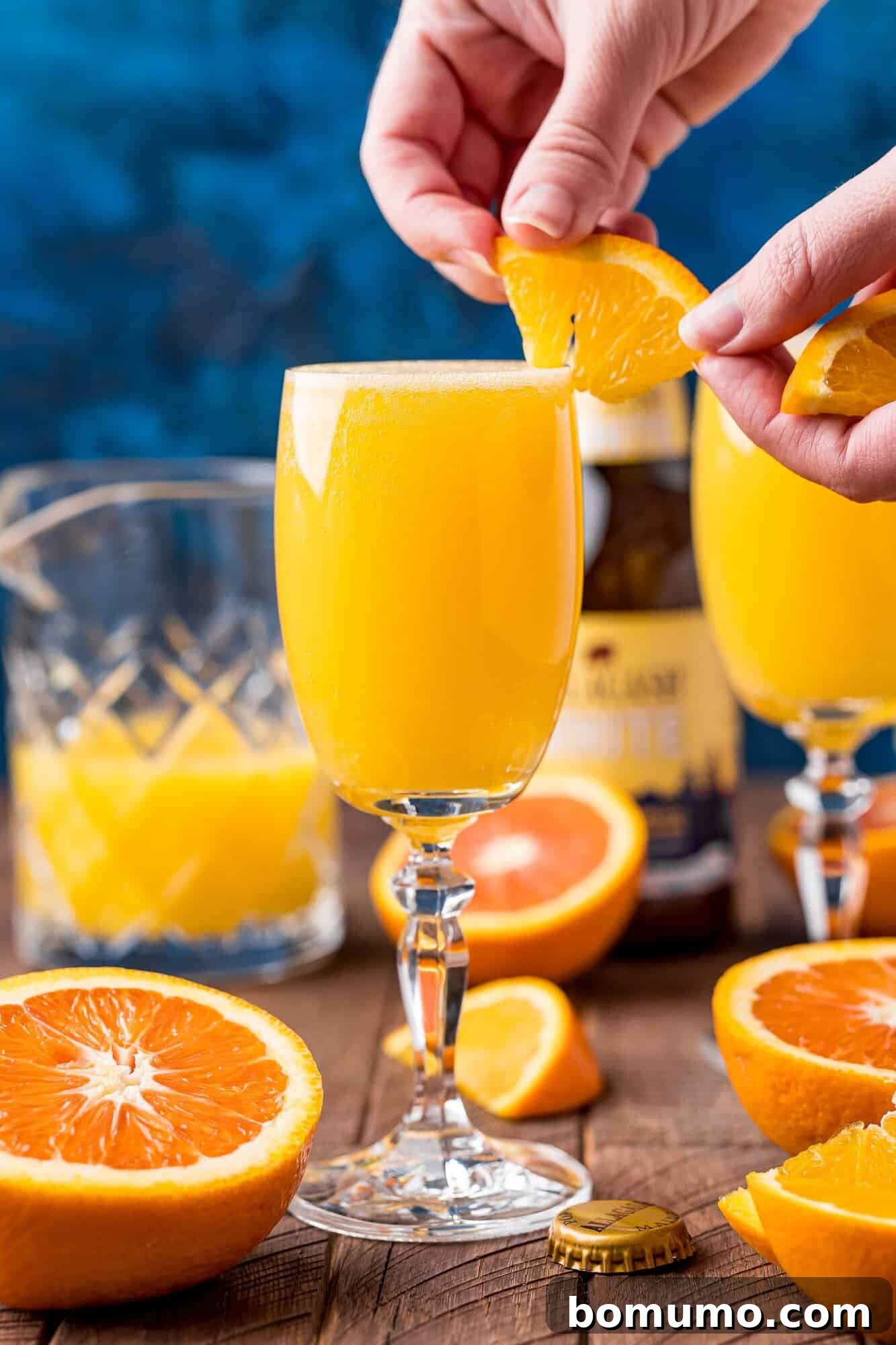 A hand delicately places a fresh orange slice as a garnish onto a tall, amber-colored Beermosa cocktail. The glass sits on a wooden table, surrounded by more fresh oranges and a beer bottle, completing the presentation of the drink.