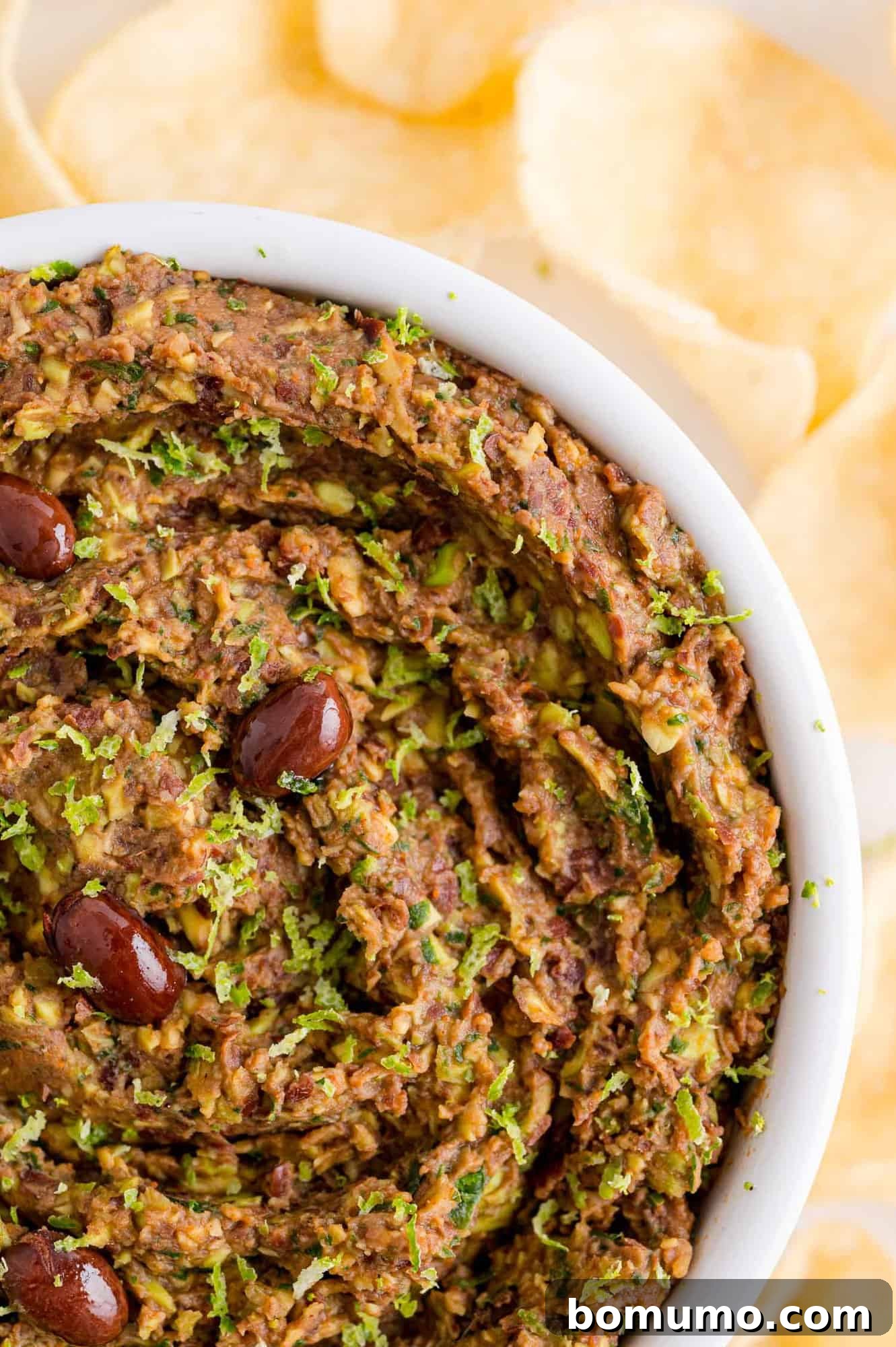 A pristine white bowl filled with creamy black bean dip, artfully arranged with a generous scattering of tortilla chips around it.