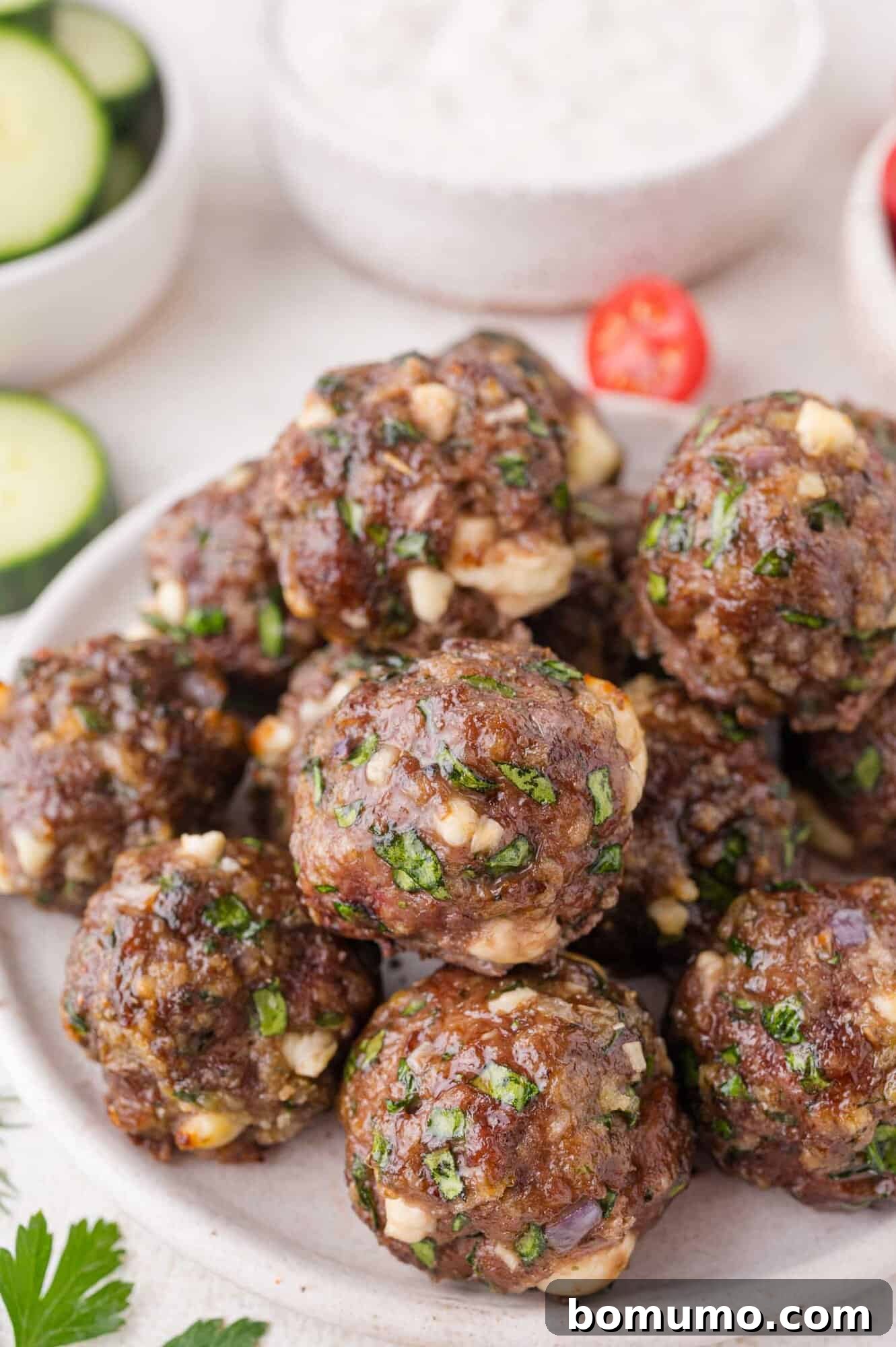 A plate of cooked Greek meatballs mixed with spinach and cheese, with bowls of sliced cucumber and sauce in the background.