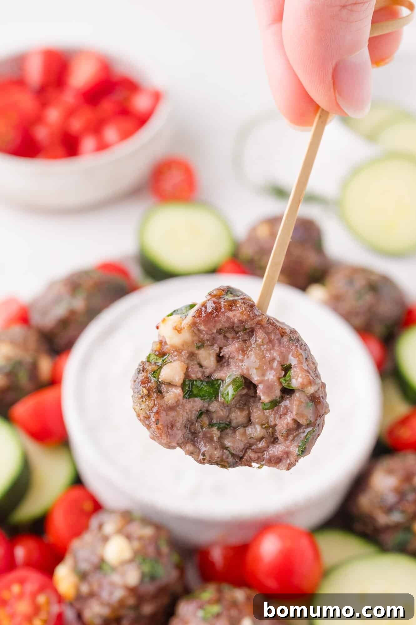 A hand holds a Greek meatball on a skewer above a bowl of white dipping sauce, surrounded by sliced cucumbers and cherry tomatoes.