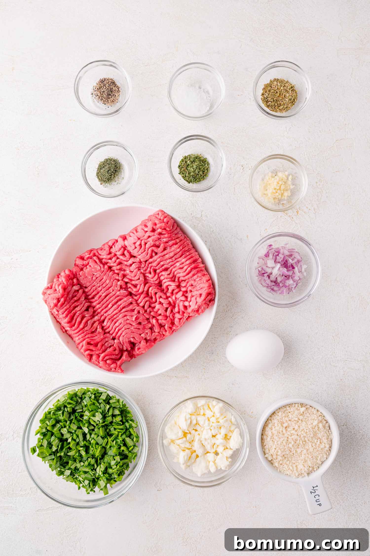 Raw ground meat, chopped spinach, diced onion, minced garlic, feta cheese, egg, breadcrumbs, and assorted seasonings are arranged in separate bowls on a white surface.