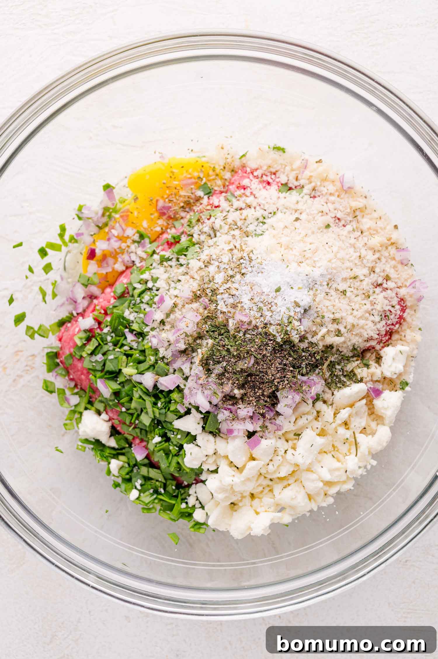 A glass bowl containing chopped spinach, onions, crumbled cheese, ground meat, an egg, breadcrumbs, and various seasonings, ready to be mixed.